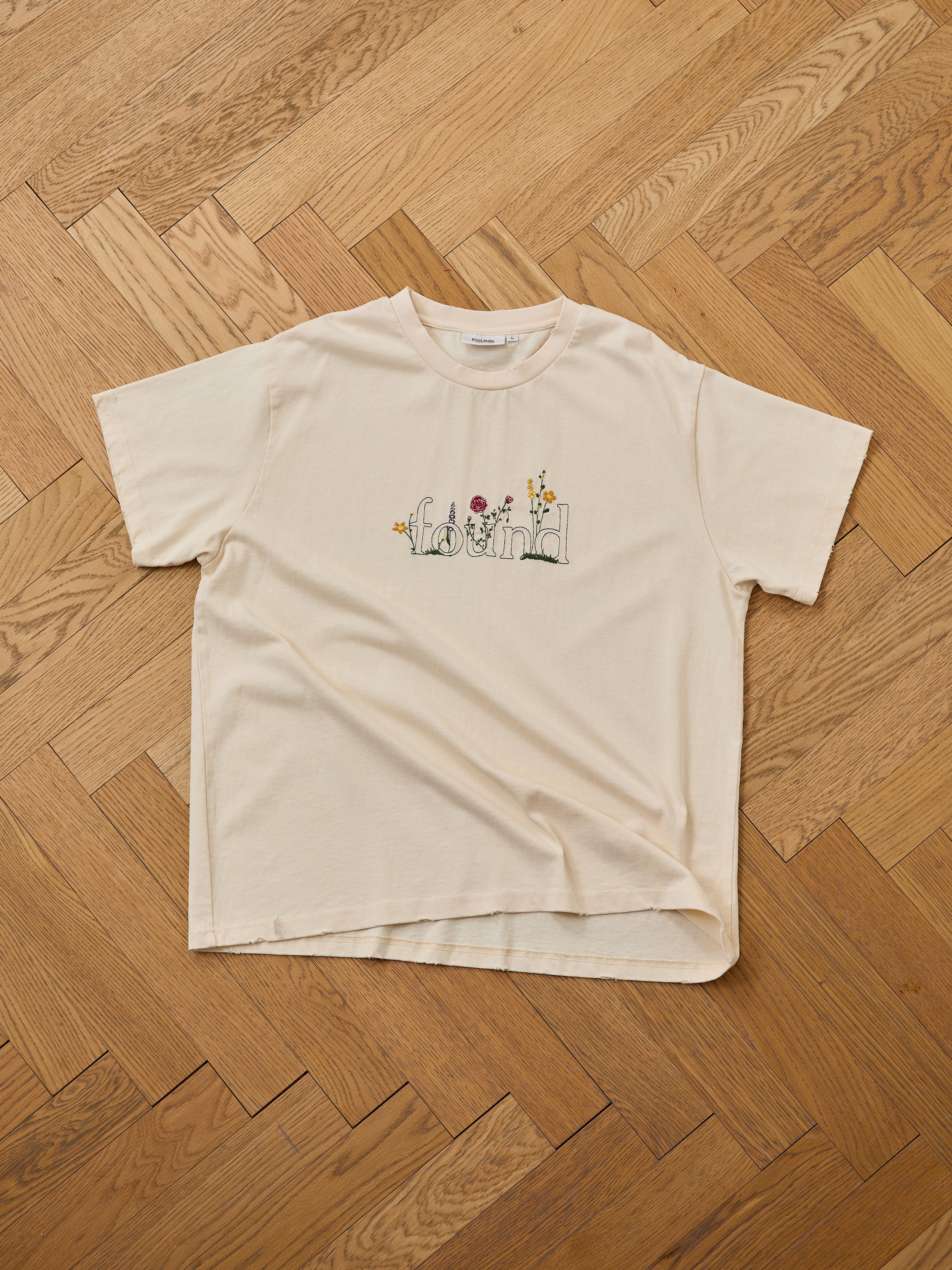 The FOUND Found Logo Flower Tee, in beige with short sleeves, features "found" and embroidered flowers for a subtle vintage look—shown laid flat on a wooden herringbone floor.