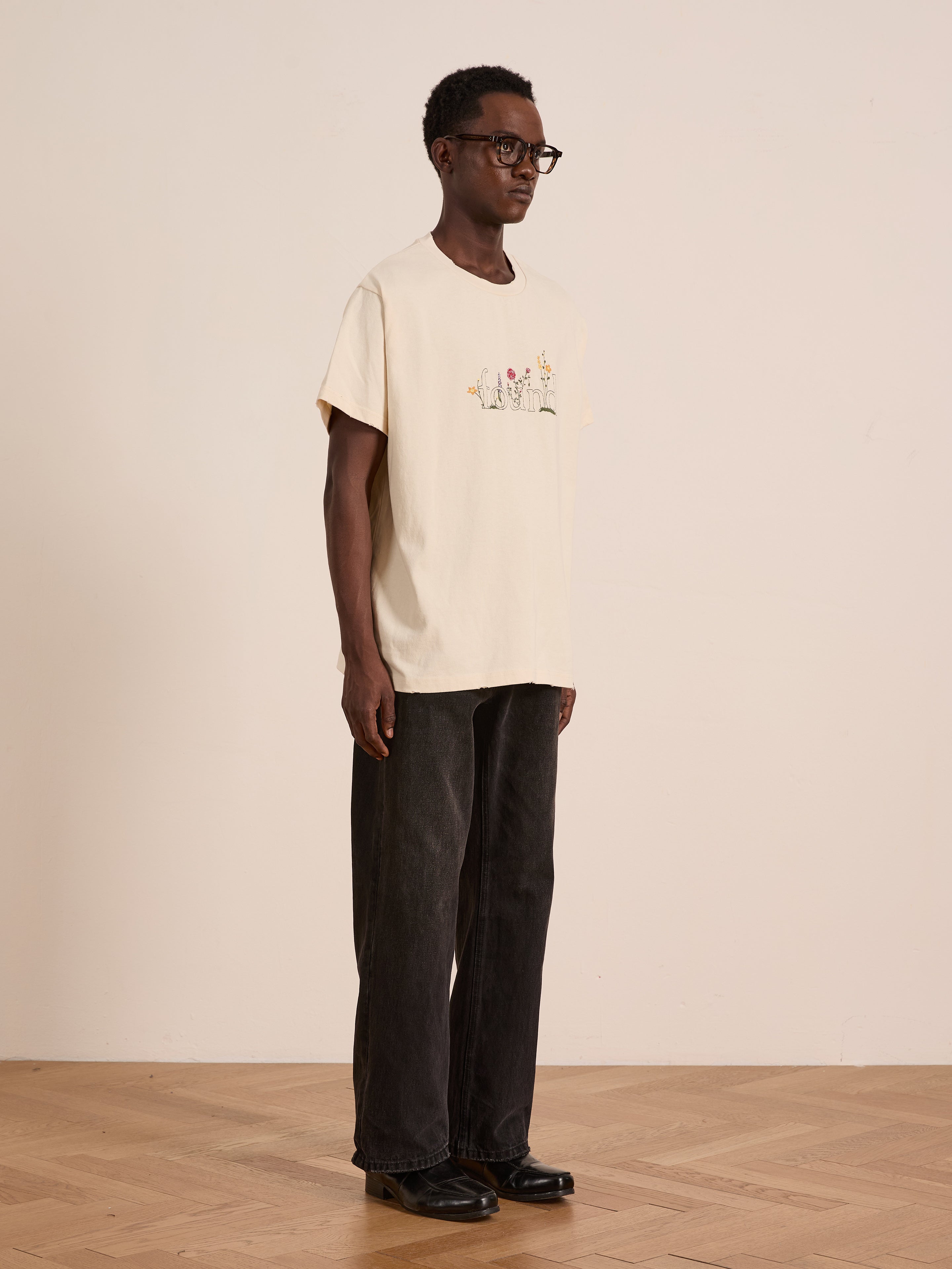 A person wearing glasses, a light-colored FOUND Found Logo Flower Tee, black wide-leg pants, and black shoes stands indoors on a wooden floor against a plain wall, giving their look a subtle vintage touch.
