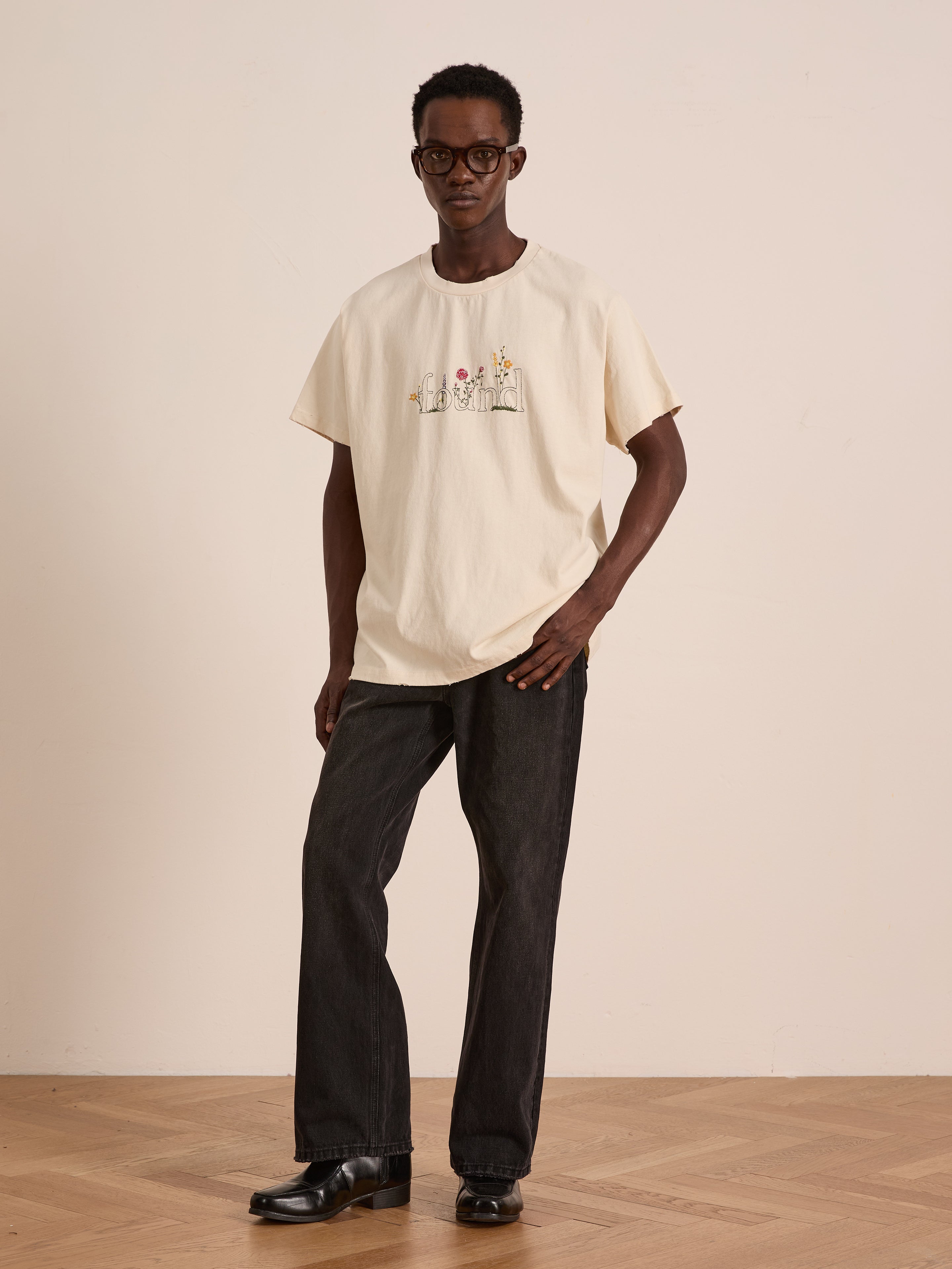 A man wearing glasses stands in a neutral room, dressed in a cream-colored FOUND Found Logo Flower Tee with embroidered blooms, paired with black jeans and black shoes.