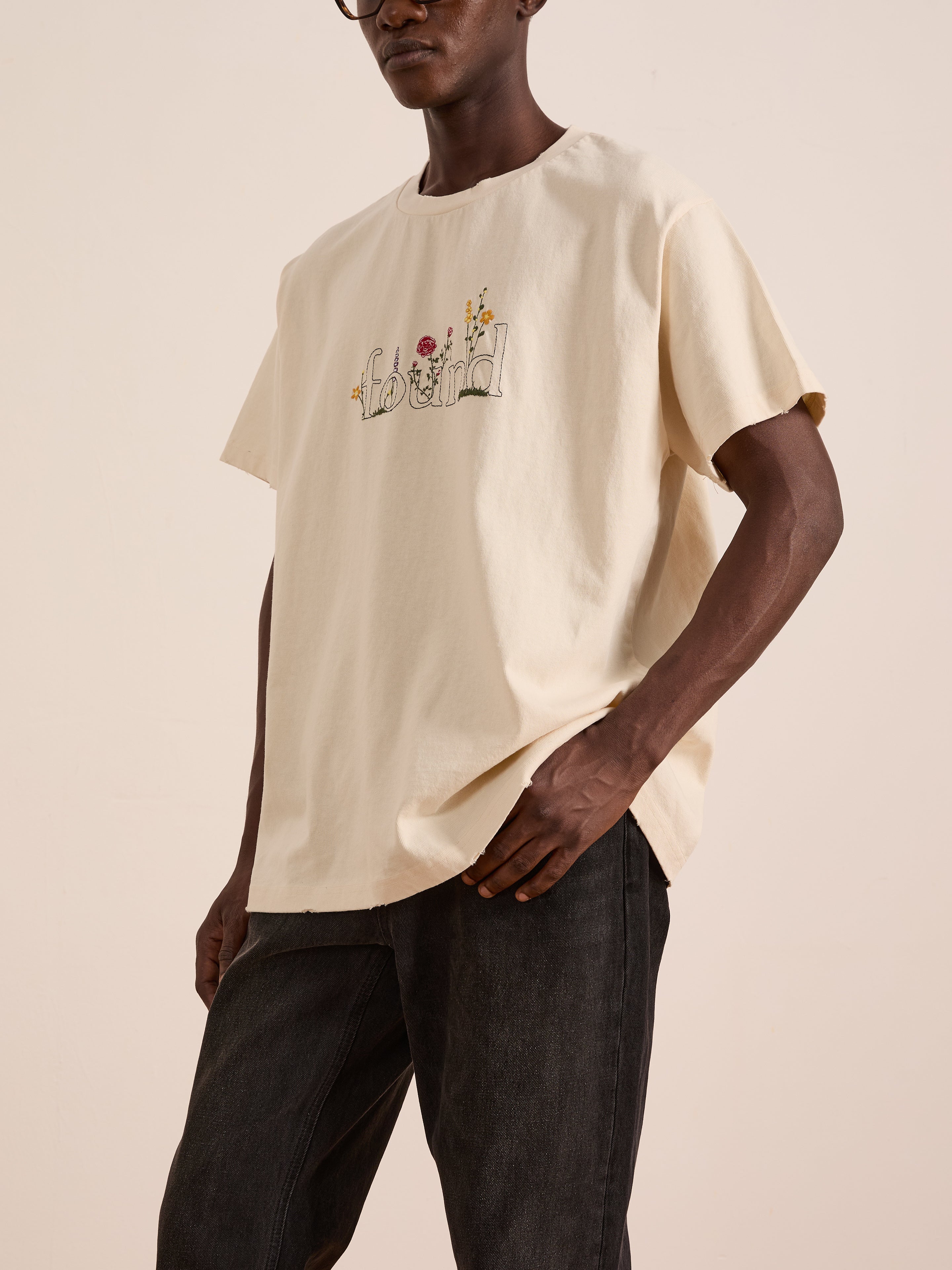 A person wearing the FOUND Found Logo Flower Tee—a loose, cream cotton top with floral embroidery and “bloom” text—paired with dark pants.