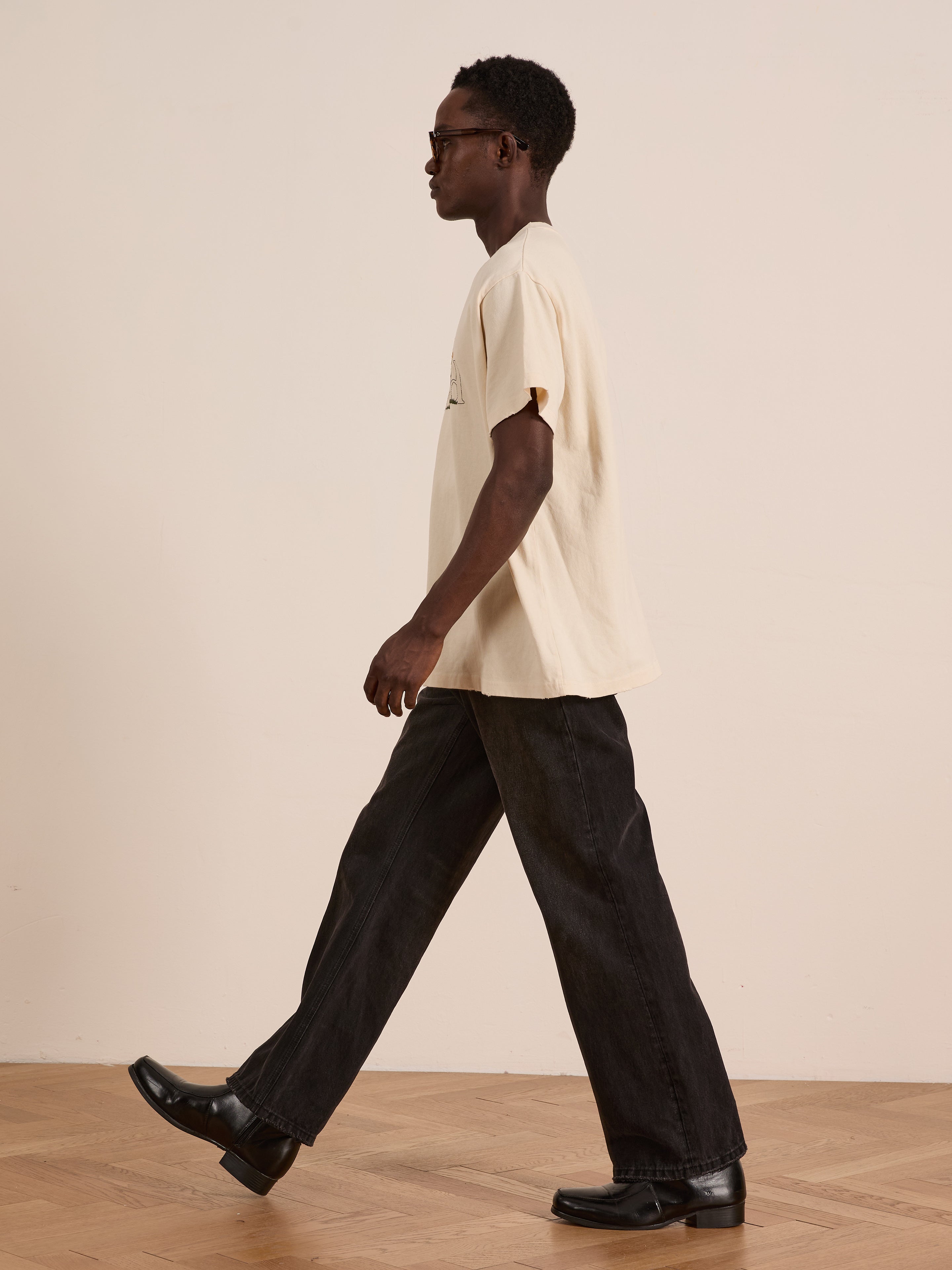 Wearing the FOUND Found Logo Flower Tee with embroidered blooms, black jeans, and black shoes, a person walks indoors on a wooden floor against a plain wall, giving their outfit a vintage vibe.