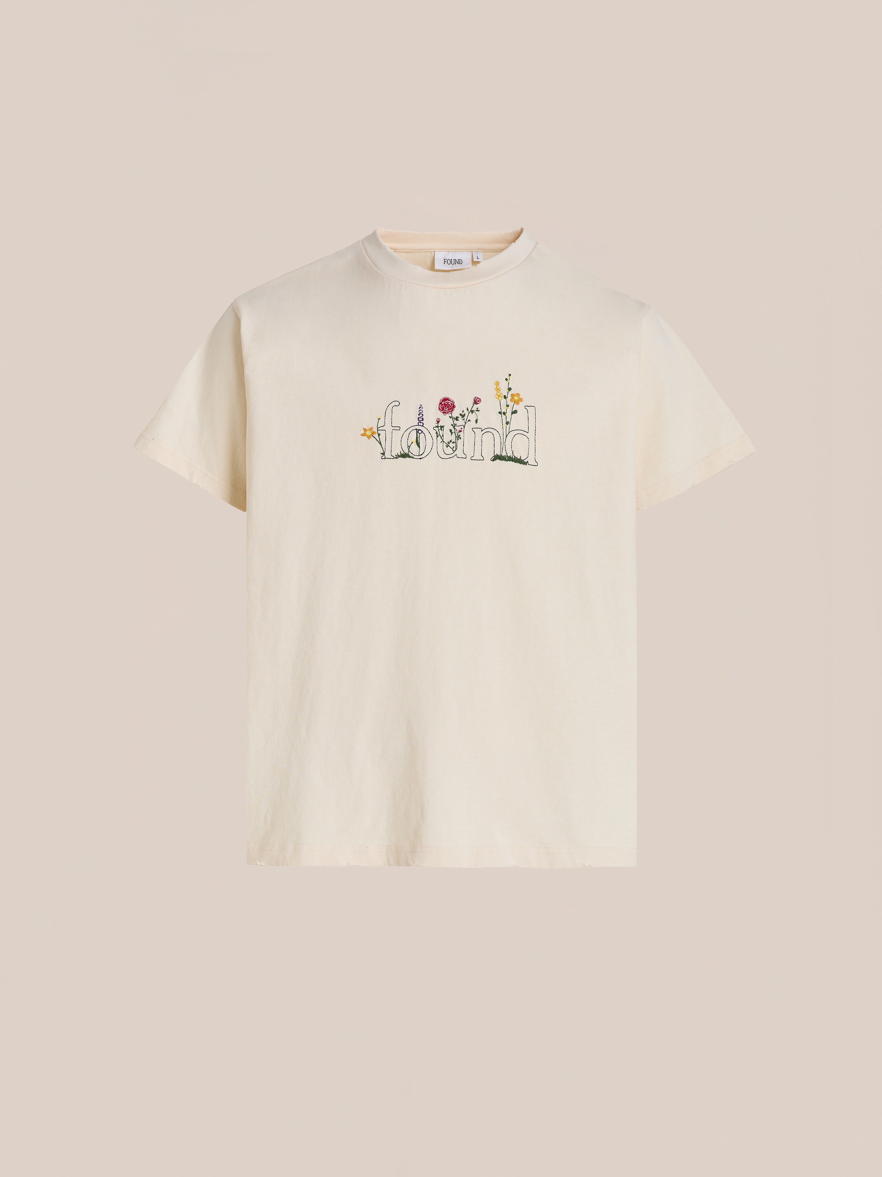 The FOUND Found Logo Flower Tee is cream-colored with “found” embroidered on the chest, featuring delicate blooms and small colorful flowers for a vintage-inspired look.