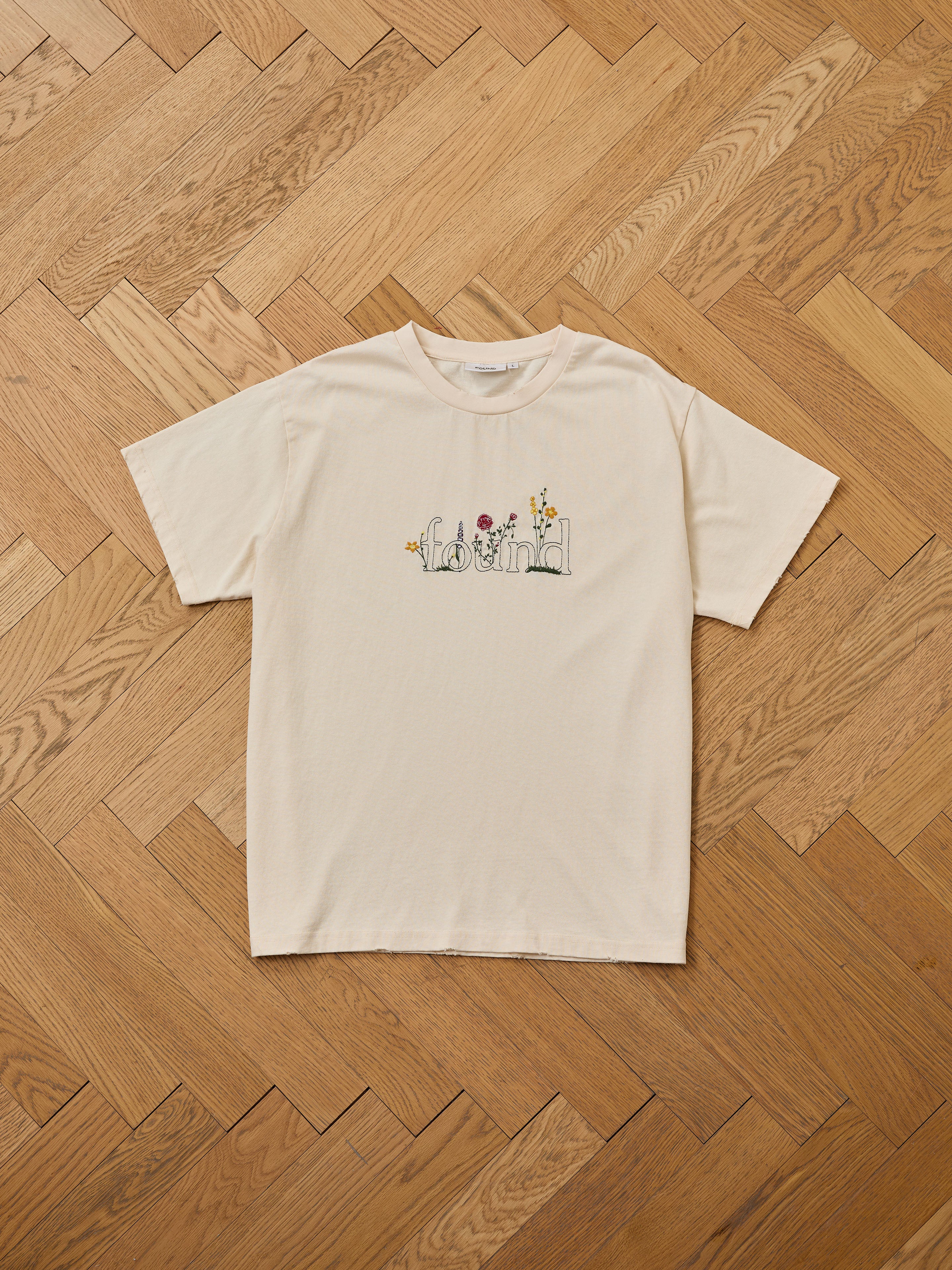 The FOUND Found Logo Flower Tee features "found" and embroidered blooms on the front for a vintage vibe. This beige tee, decorated with small flowers, is displayed flat on a wooden herringbone-patterned floor.