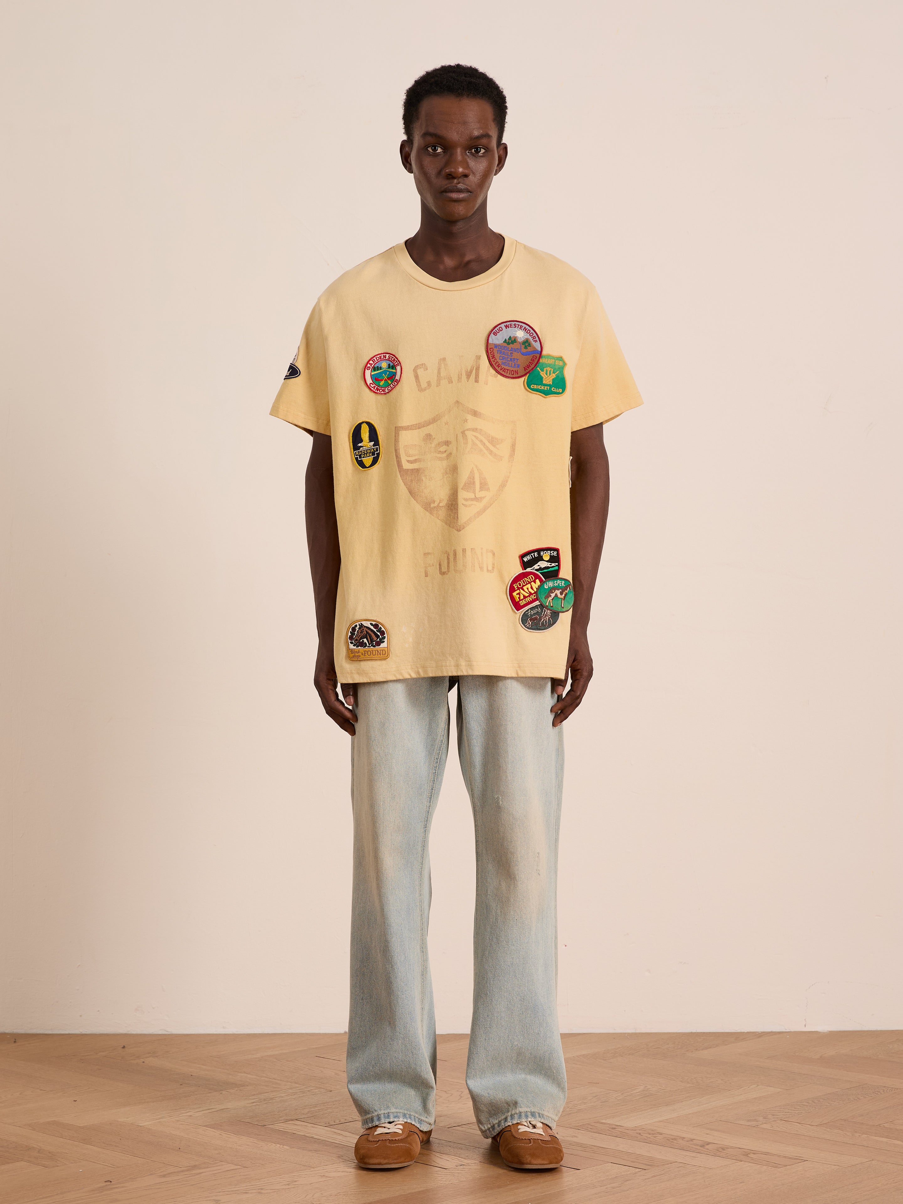 Indoors against a plain wall, a person wears the FOUND Patchwork Camp Tee—unisex, relaxed fit, and finished with vintage-inspired embroidered patches—styled with light blue jeans and brown shoes.