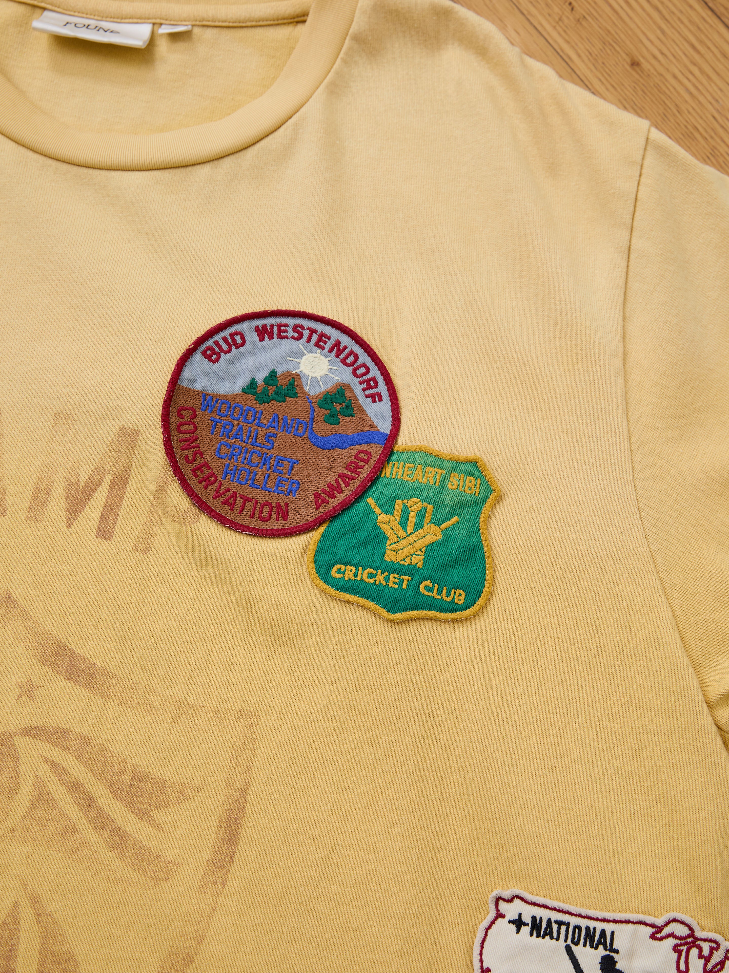 The FOUND Patchwork Camp Tee in yellow features vintage-style embroidered patches with "Bud Westendorf Woodland Trails Conservation Award" and "Heart Sibi Cricket Club" text, offered in a unisex relaxed fit.