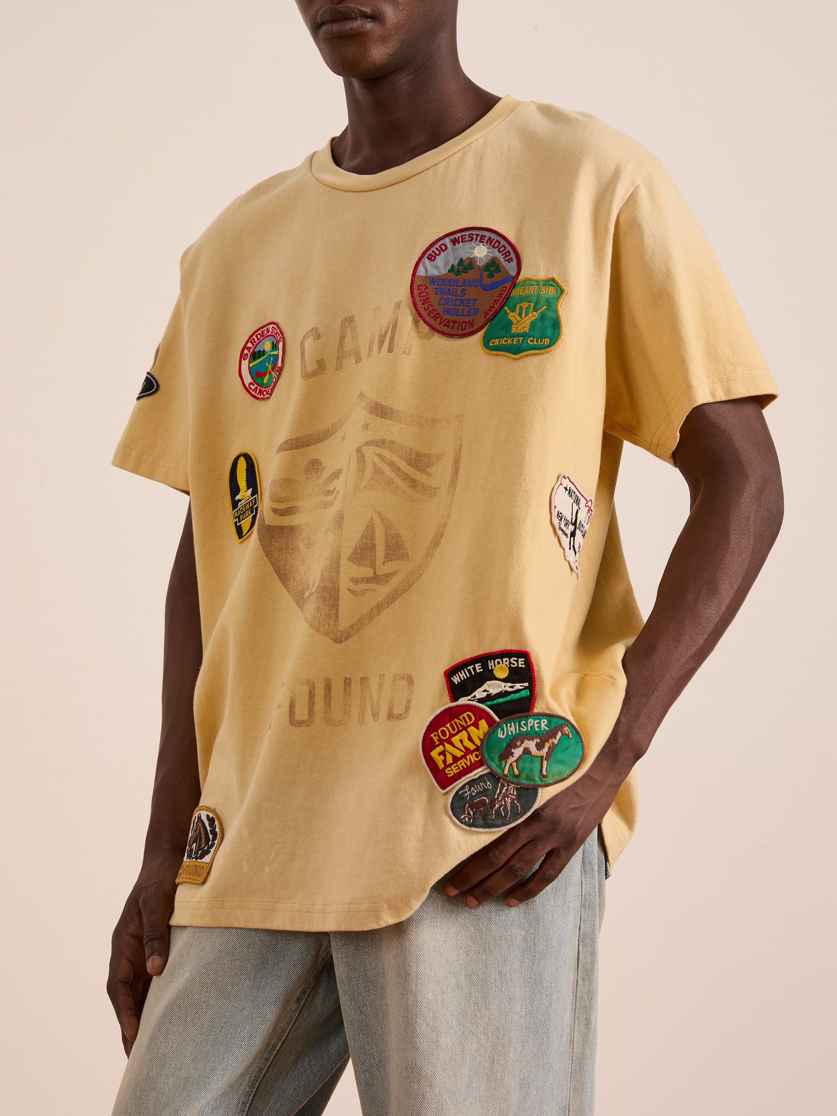 Someone in a FOUND Patchwork Camp Tee—unisex, relaxed fit, yellow with vintage-style embroidered patches—paired with light blue jeans.