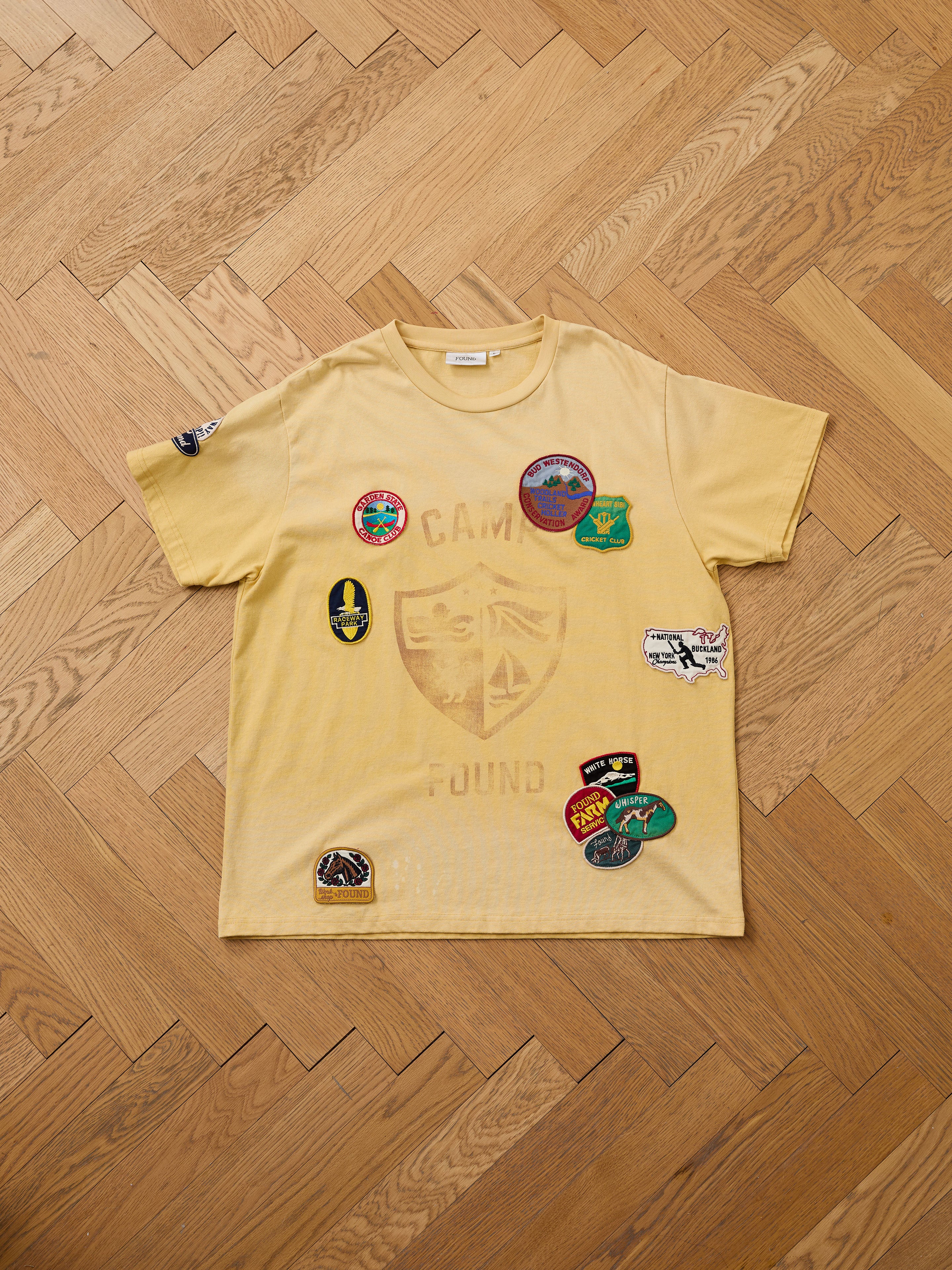 The FOUND Patchwork Camp Tee in yellow, featuring a faded "Camp Found" emblem and vintage-style embroidered patches, is laid flat on a herringbone floor to highlight its unisex relaxed fit.