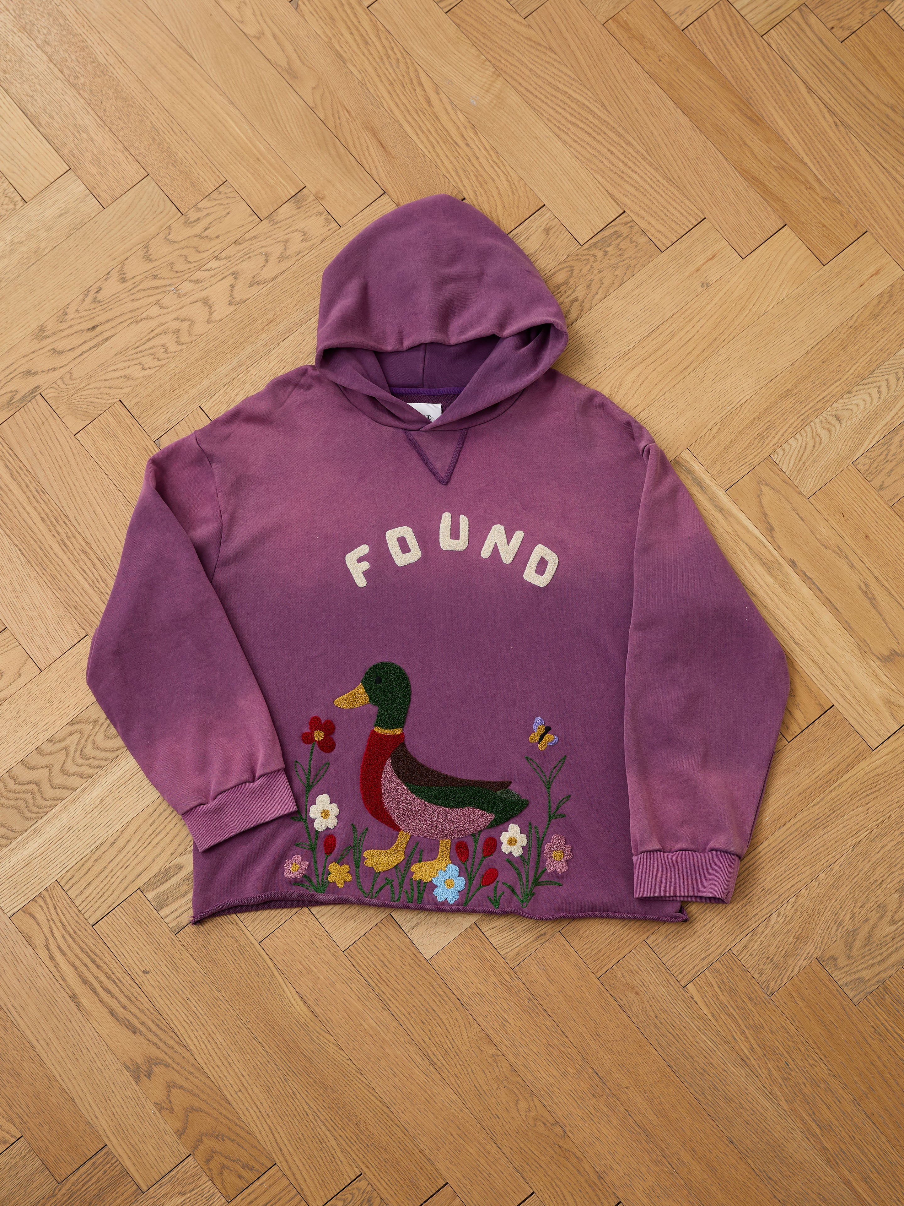 The FOUND Embroidered Wildlife Hoodie, featuring a purple design with "FOUND" on the chest and an embroidered duck with flowers along the hem, is crafted from soft vintage French-terry and shown laid flat on a wooden floor.