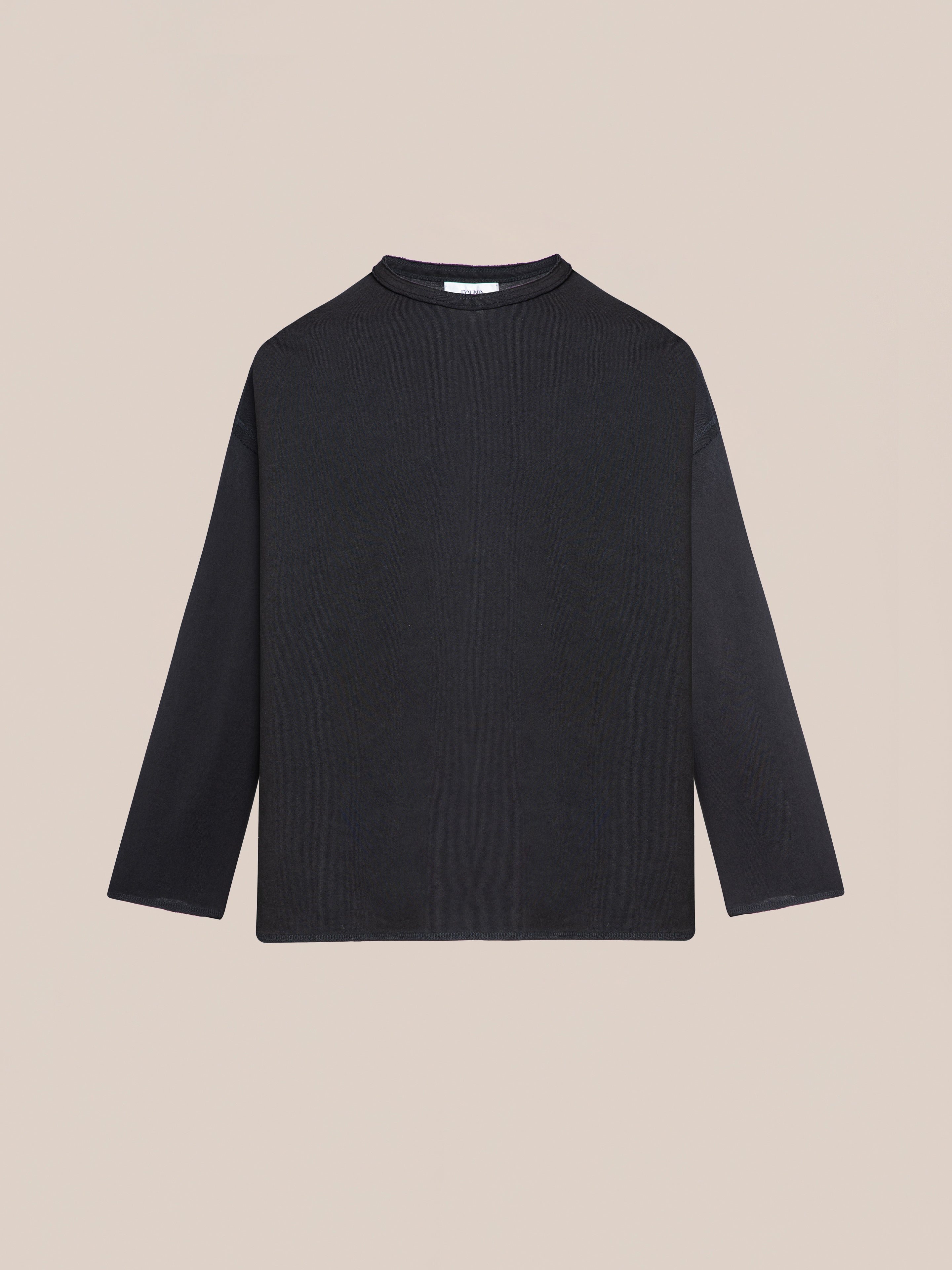 The FOUND Reversed LS Tee in plain black, featuring a relaxed fit and crew neckline, is displayed against a beige background.