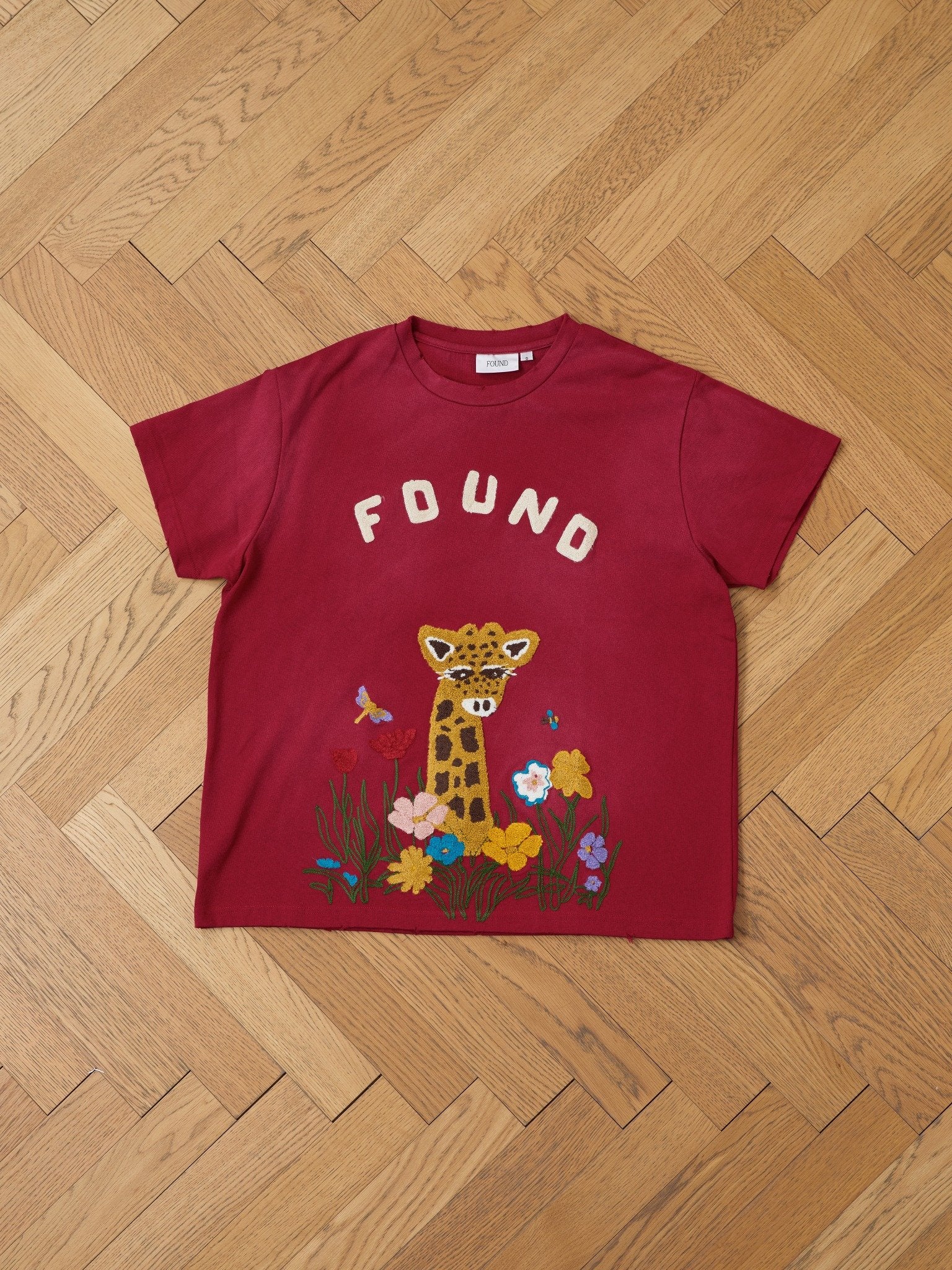 The FOUND Embroidered Wildlife Tee is a red unisex relaxed fit shirt featuring "FOUND" on the chest and chenille embroidery of a giraffe, flowers, and butterflies near the hem, displayed flat on a wooden herringbone floor.