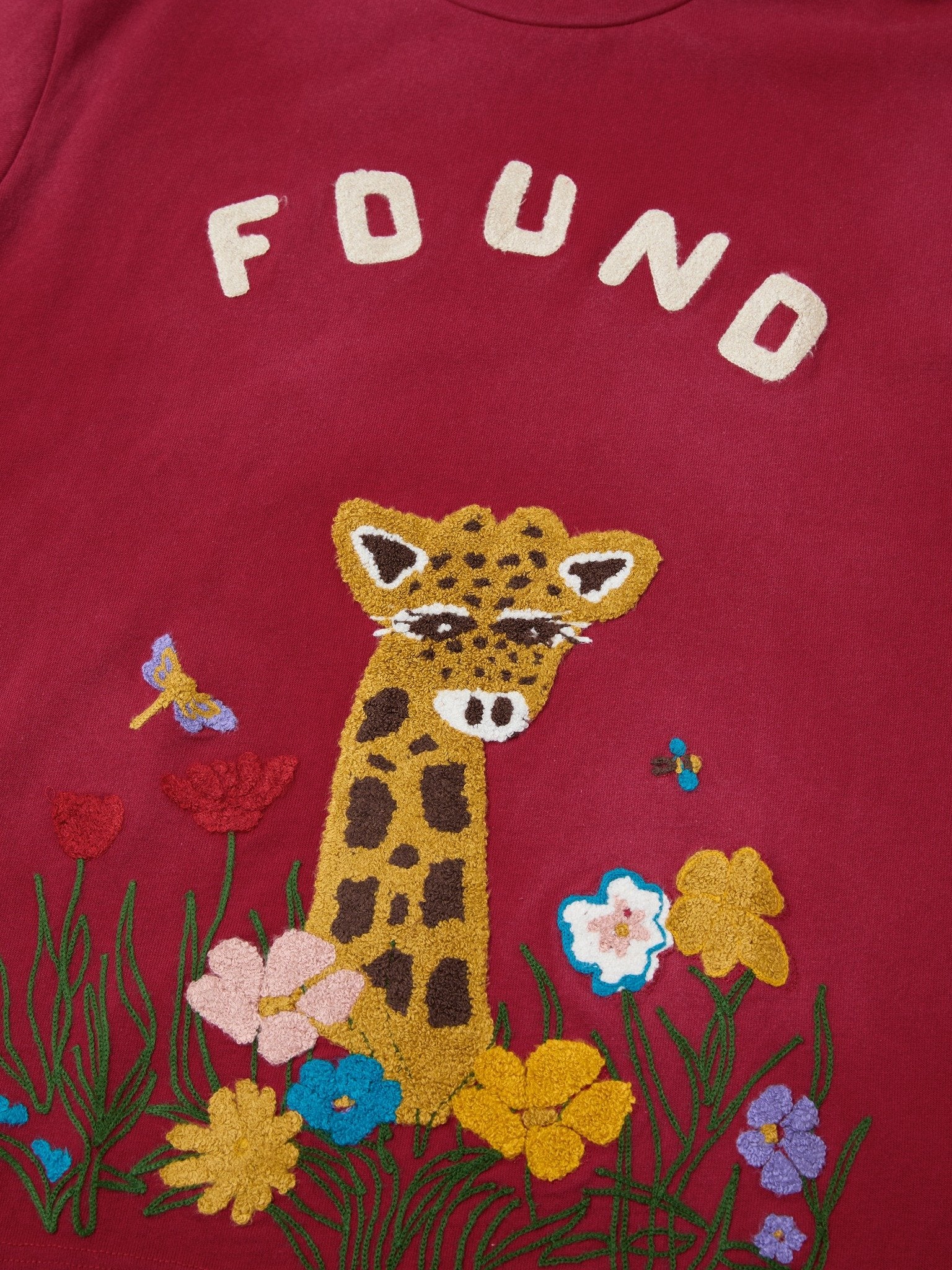 The FOUND Embroidered Wildlife Tee features a maroon color, a chenille giraffe head surrounded by colorful flowers and butterflies, and "FOUND" lettering. Designed in a unisex relaxed fit for everyday comfort.