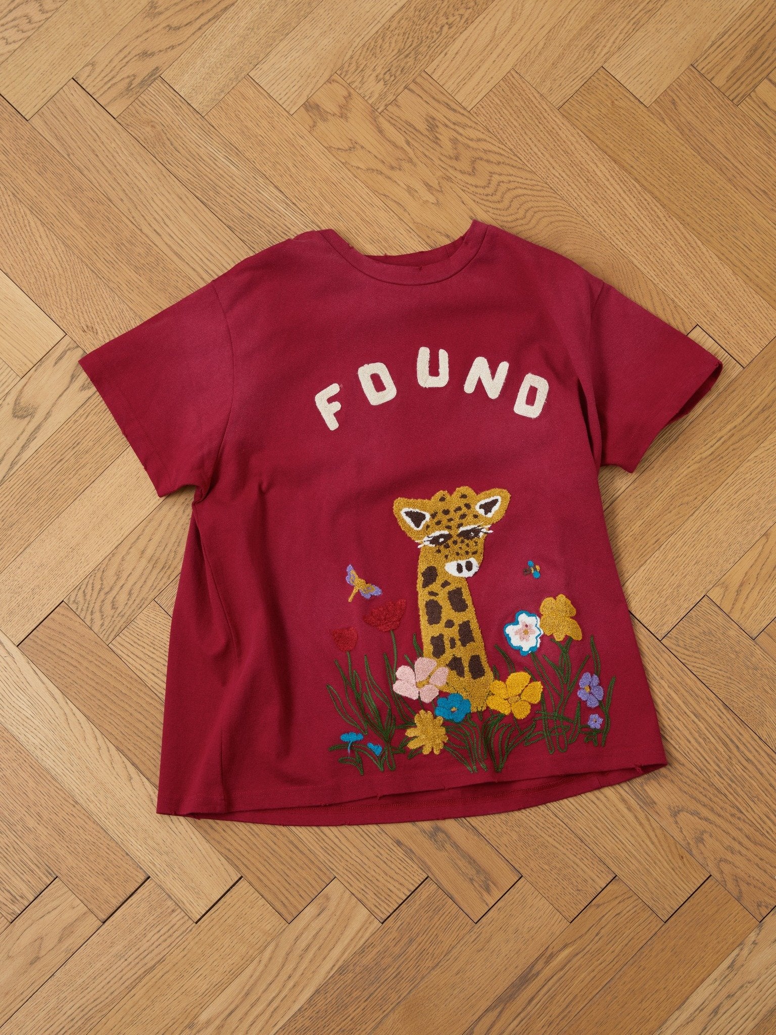 The FOUND Embroidered Wildlife Tee in red features chenille embroidery with "FOUND" above a giraffe and colorful flowers. Unisex relaxed fit for everyday comfort. Shown laid flat on a wooden herringbone floor.