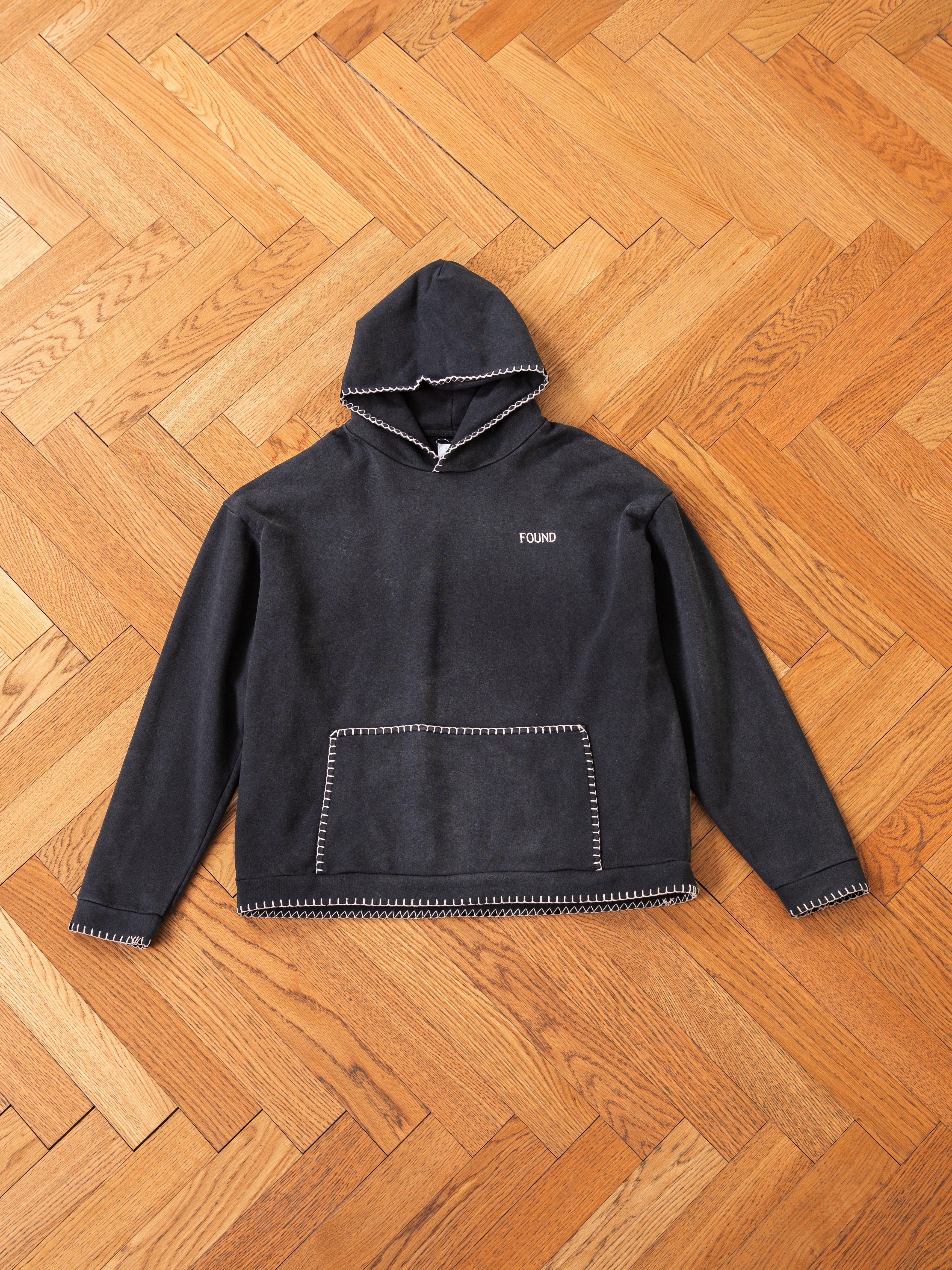 The Stitched Border Distressed Logo Hoodie from FOUND, a vintage black hooded sweatshirt made from French terry cotton and featuring white stitching details with the brand name "FOUND" on the chest, rests gracefully on a wooden herringbone floor.