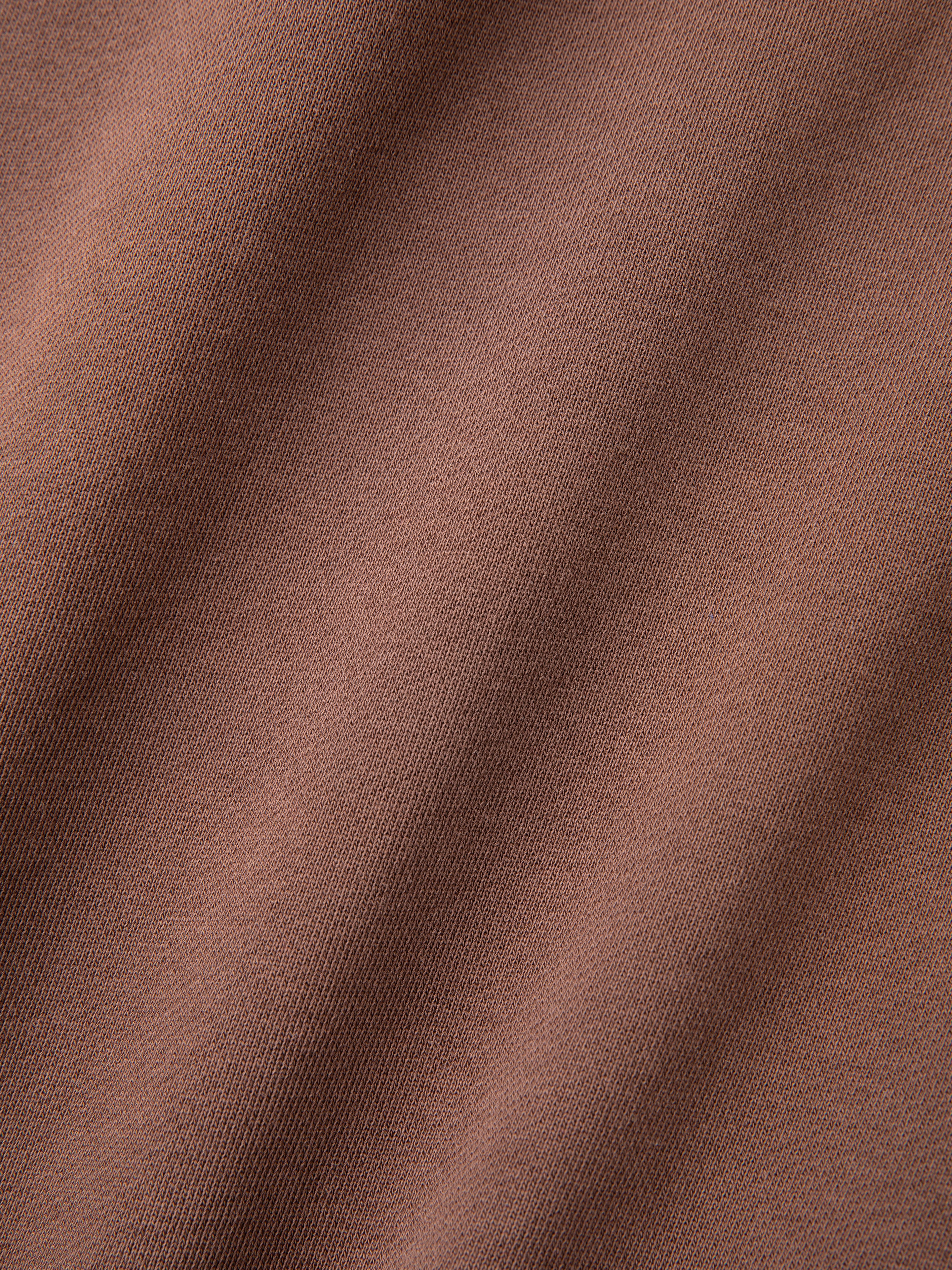 Close-up of the Patterned Logo Crewneck by FOUND, showcasing brown fabric with a smooth, slightly ribbed texture and diagonal shadows typical of French terry cotton.