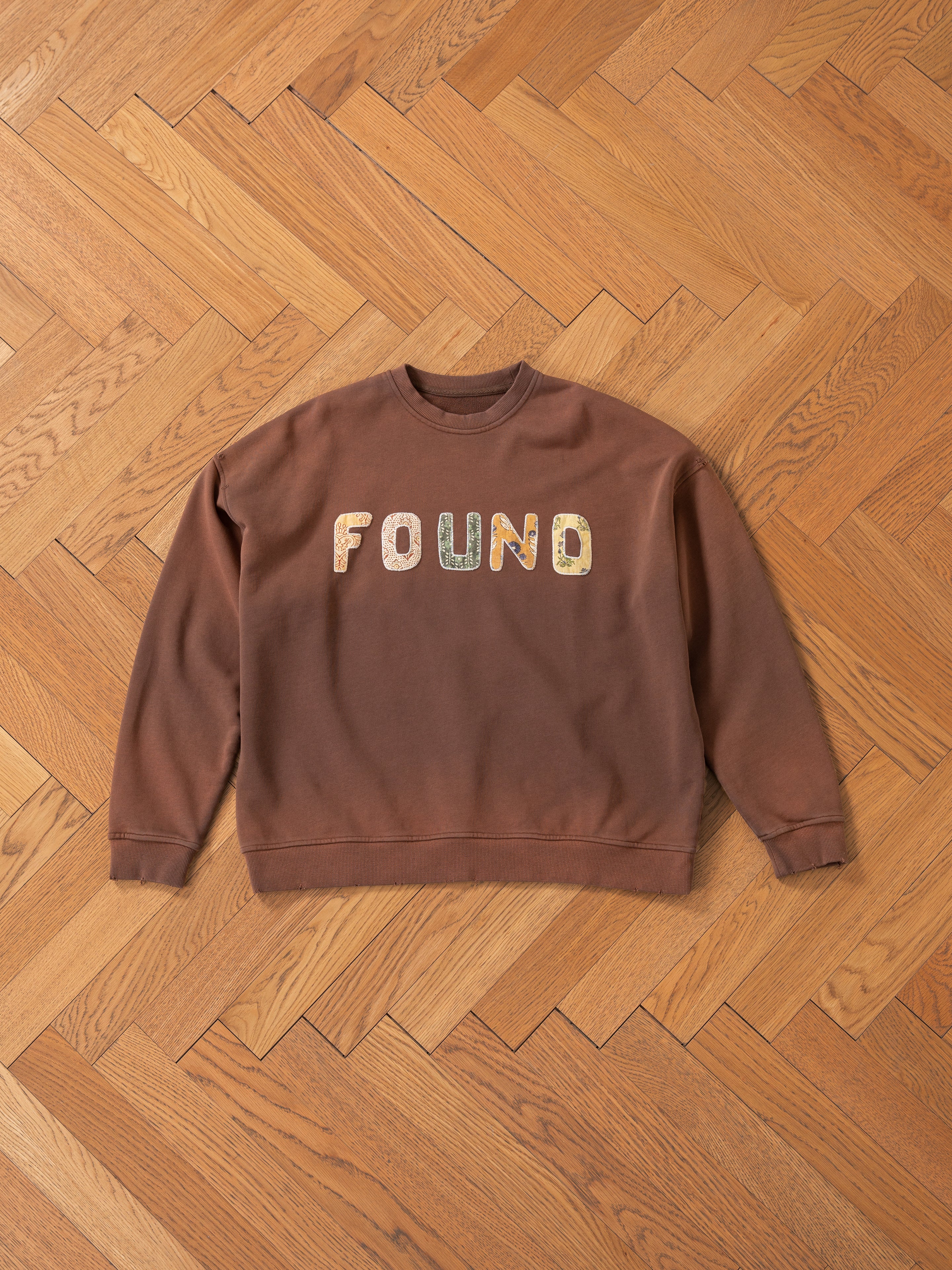 The Patterned Logo Crewneck by FOUND, made of brown French terry cotton with an appliqué "FOUND" logo, is displayed on a stylish wooden herringbone-patterned floor.