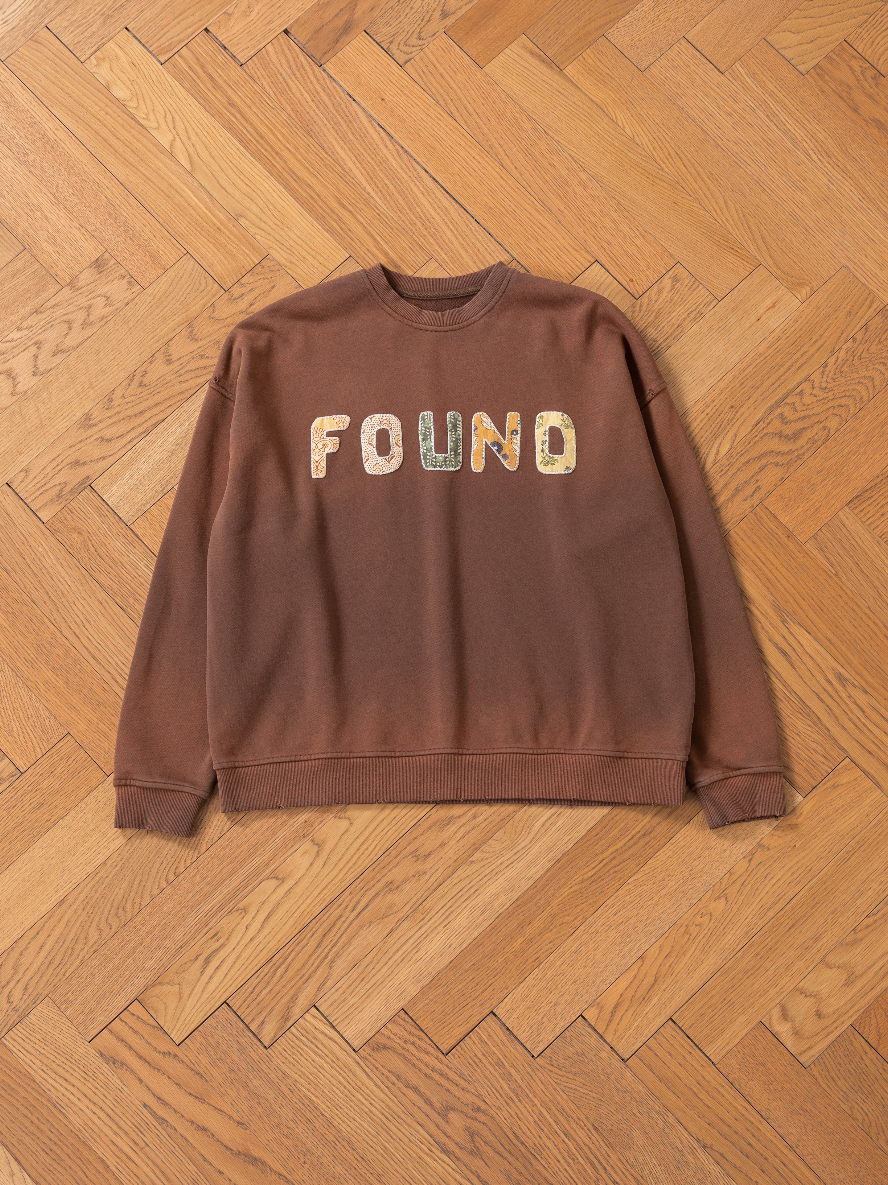 The Patterned Logo Crewneck from FOUND is a brown French terry cotton sweatshirt with multicolored "FOUND" appliqué lettering, displayed on a wooden parquet floor.