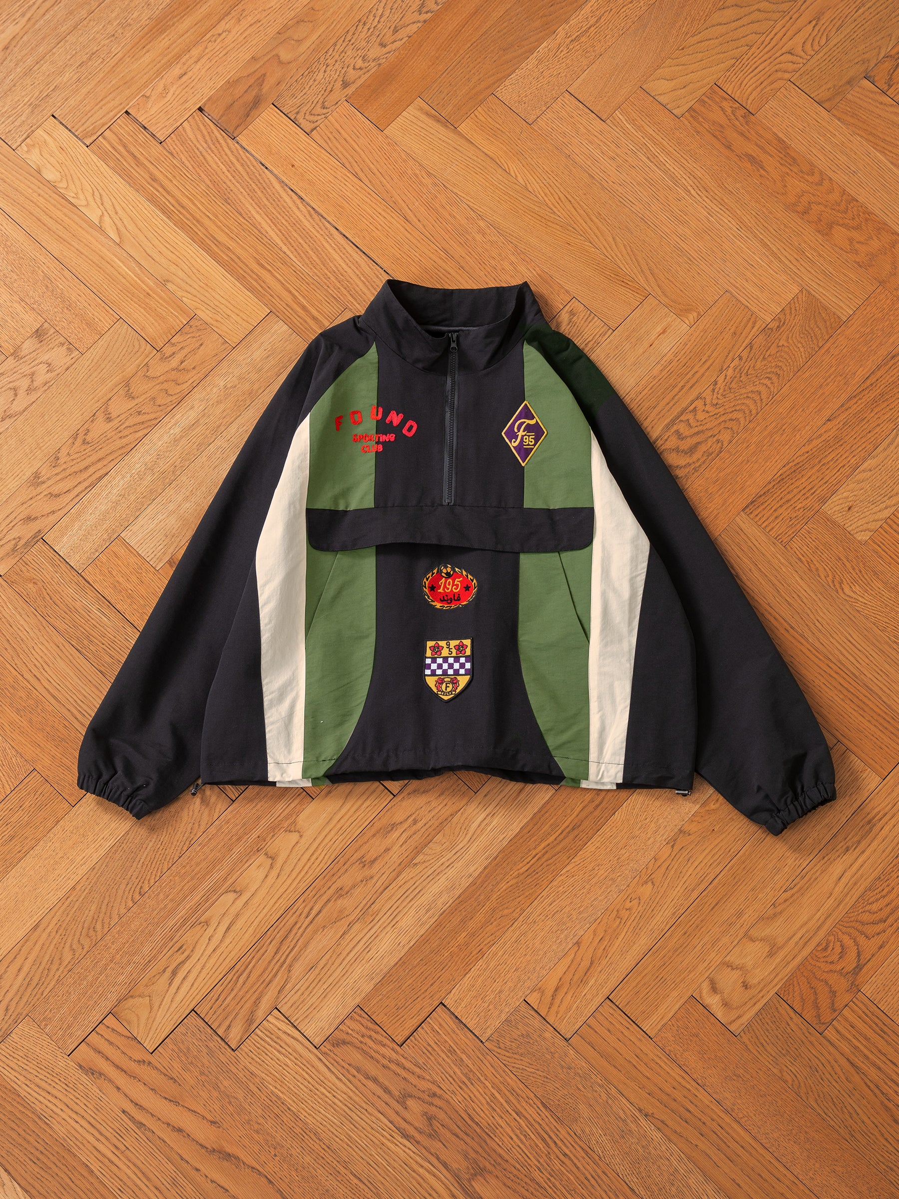 The FOUND Sporting Club Pullover Jacket, featuring a vintage aesthetic in black and green with red and white text, embroidered logos, and a high collar, is laid flat on a wooden floor.