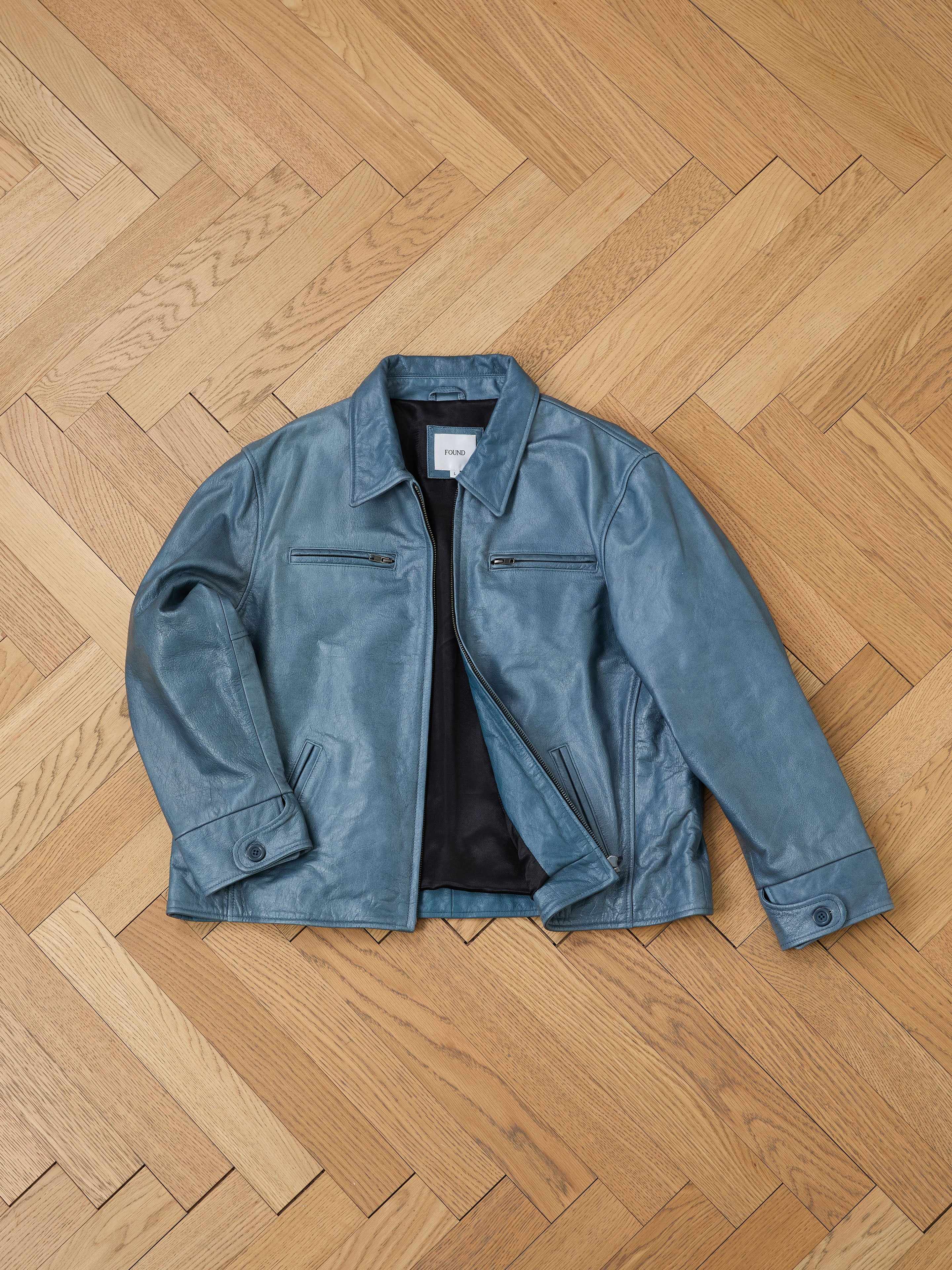 The Marlboro Sun Faded Leather Racer Jacket by FOUND lies on a wooden herringbone floor. It showcases a zippered front and chest pockets, crafted from distressed blue leather for timeless appeal with a unisex relaxed fit.