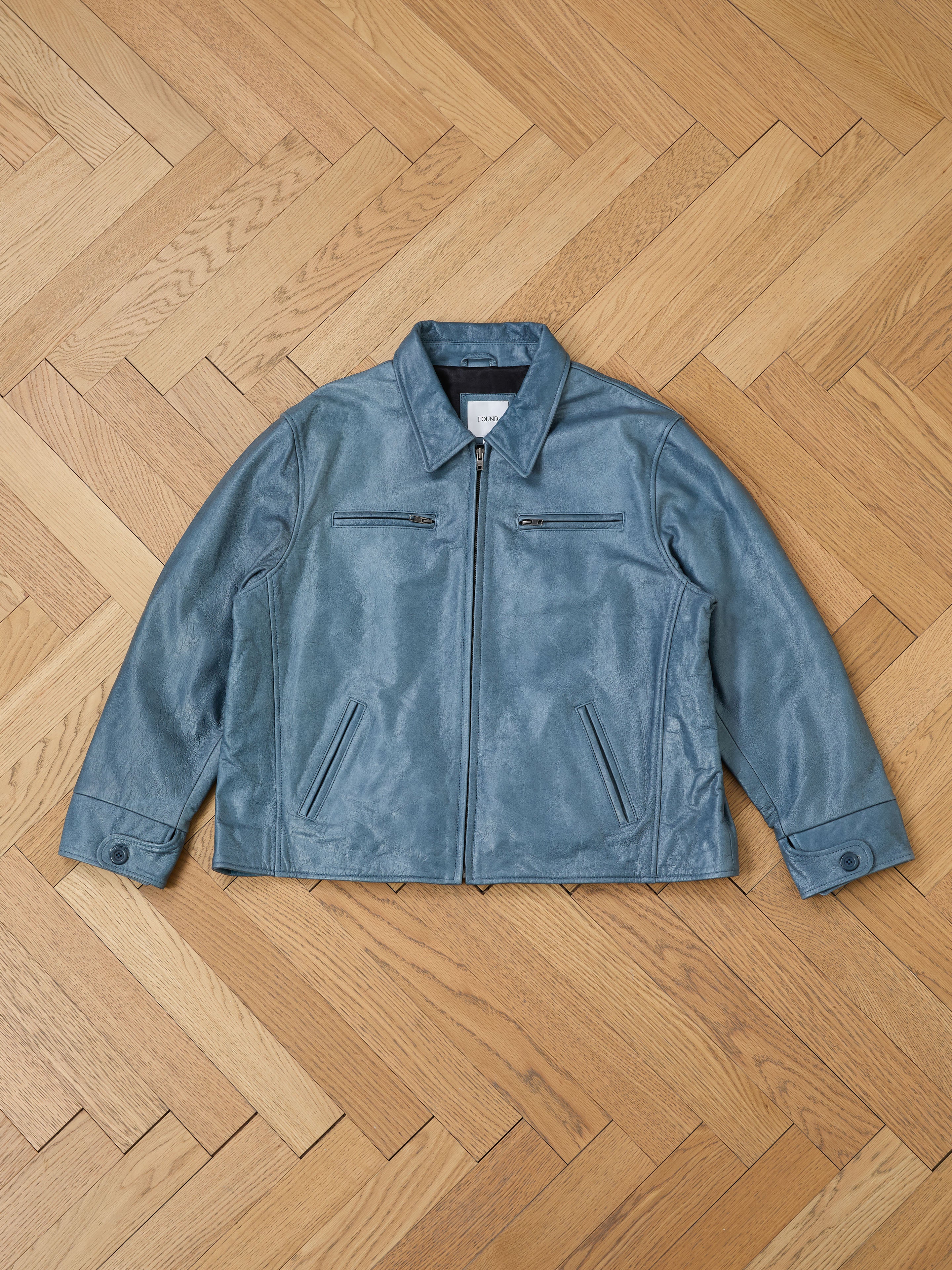 A Marlboro Sun Faded Leather Racer Jacket by FOUND, featuring a front zipper and two zippered chest pockets, lies flat on a wooden herringbone floor, highlighting its unisex relaxed fit.