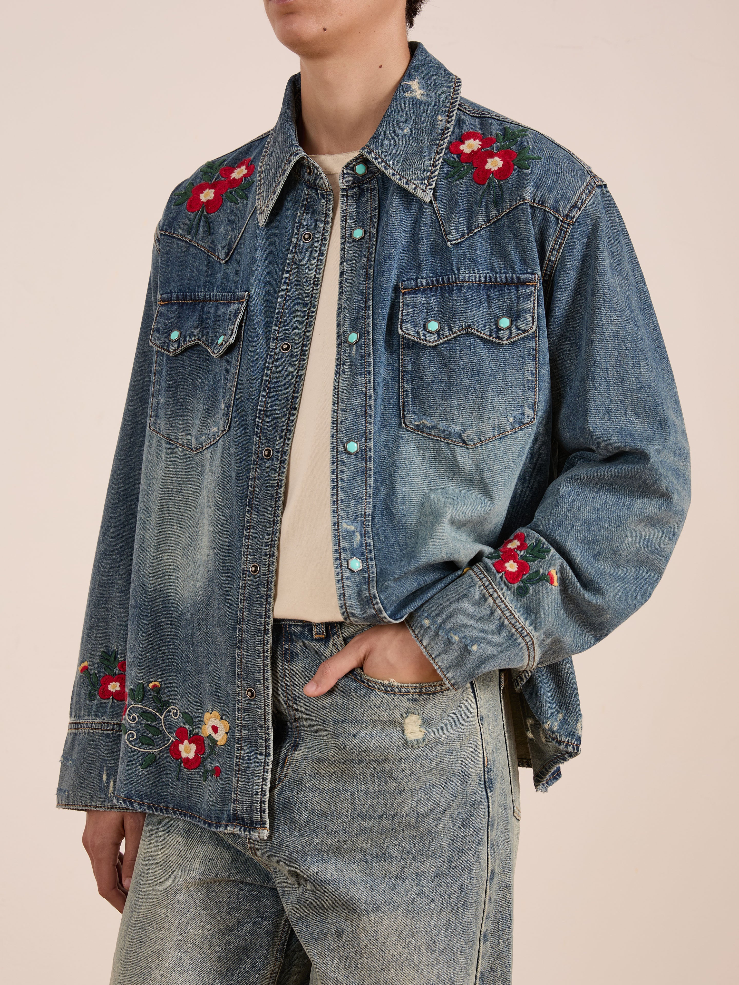 A person wears the FOUND Floral Embroidered Western Denim Shirt featuring red floral details on the shoulders, sleeves, and hem, styled with a beige t-shirt and blue jeans.