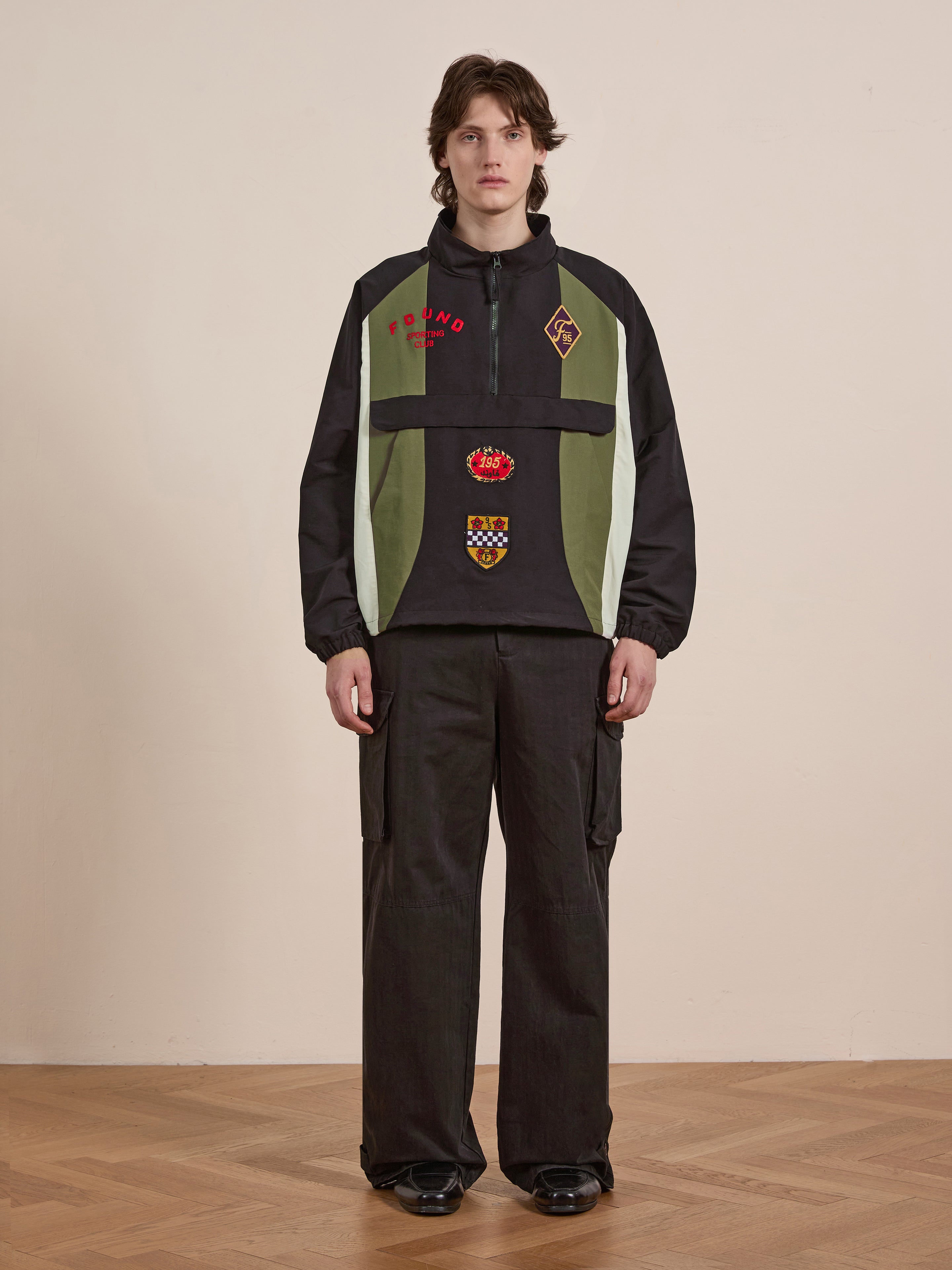 Indoors, a person exudes vintage flair in a FOUND Sporting Club Pullover Jacket in black and green with patches, alongside dark wide-leg pants and black shoes on polished wooden floors.