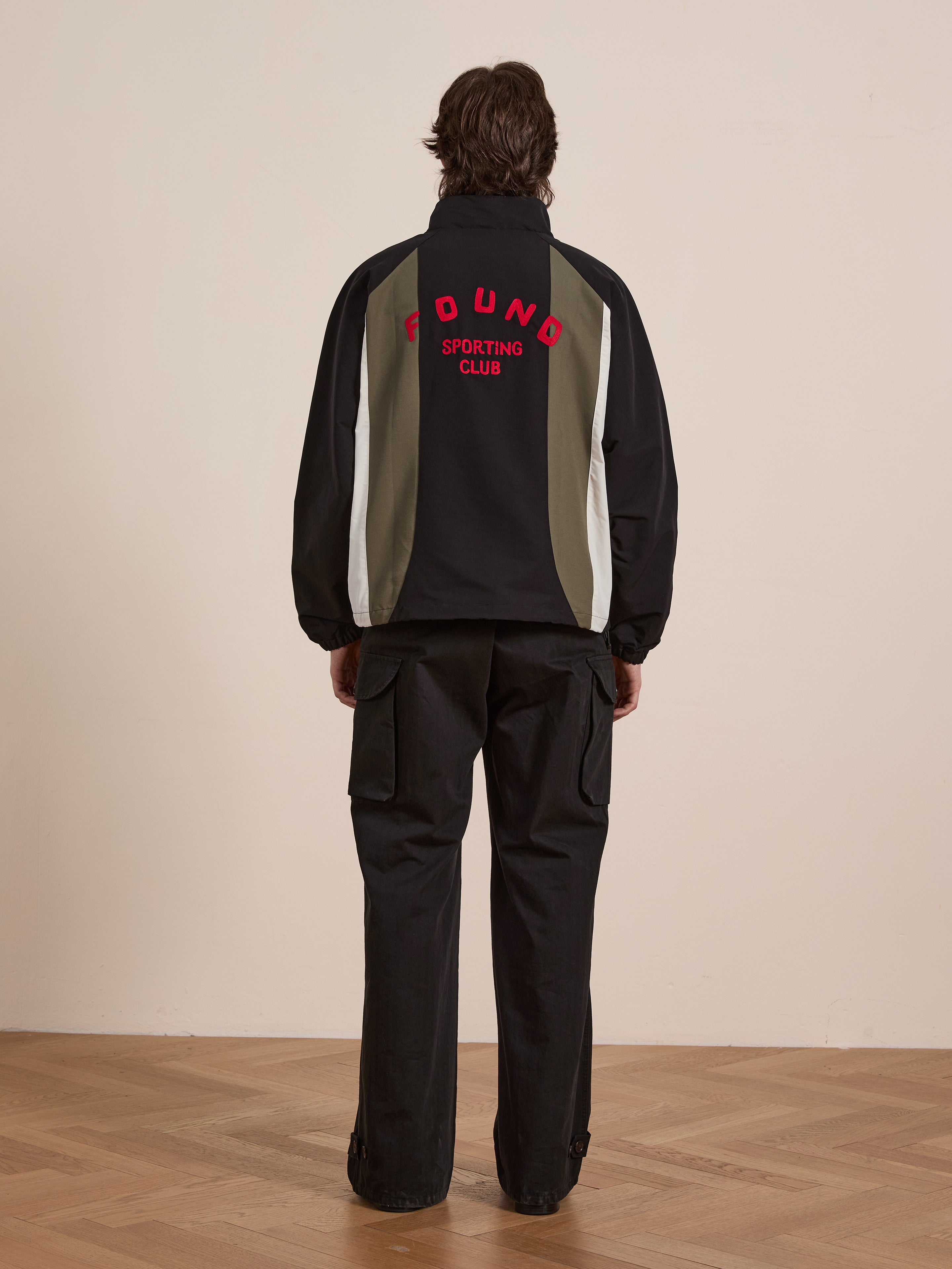 A person wearing the Sporting Club Pullover Jacket by FOUND, featuring "FOUND SPORTING CLUB" in red on a black and grey design, paired with black cargo pants, embodies a vintage aesthetic on the wooden floor. The unisex outfit enhances its timeless appeal.
