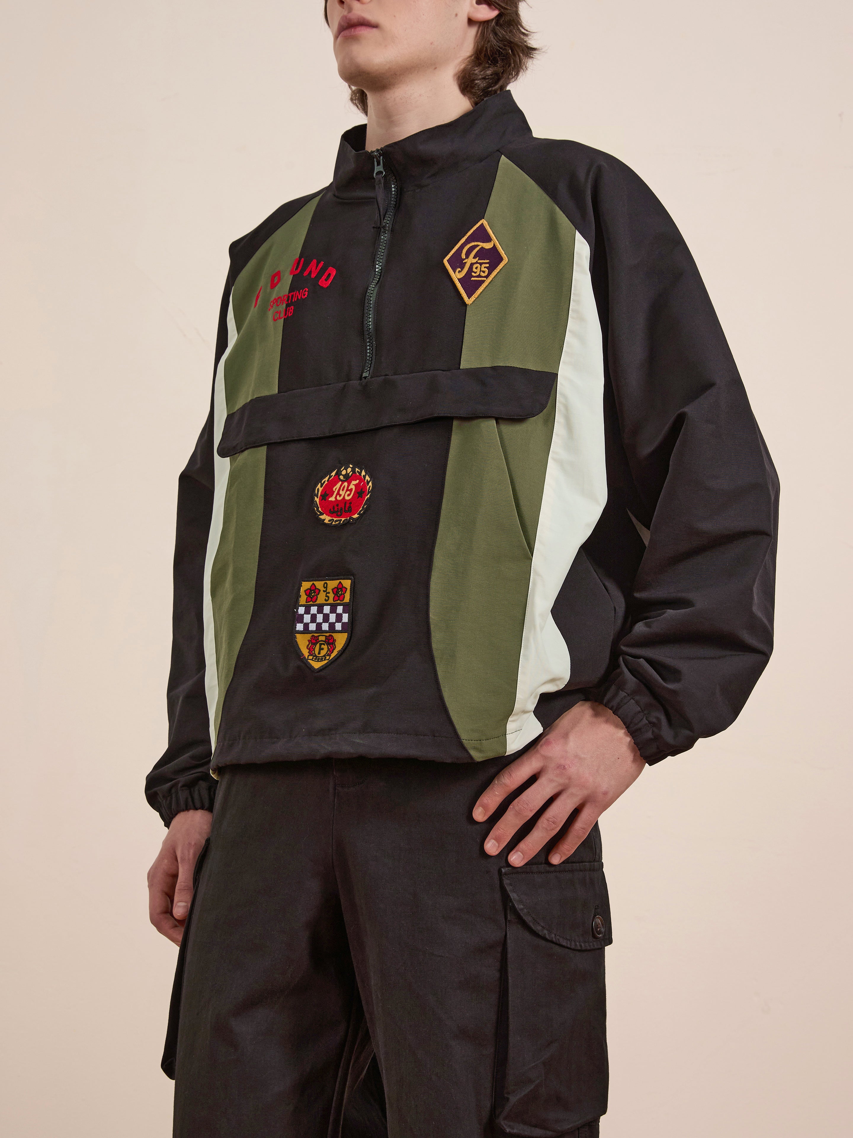 A person wearing FOUND's Sporting Club Pullover Jacket, featuring a vintage aesthetic in black, green, and white with red and multicolored patches, stands with one hand on their hip against a neutral background.