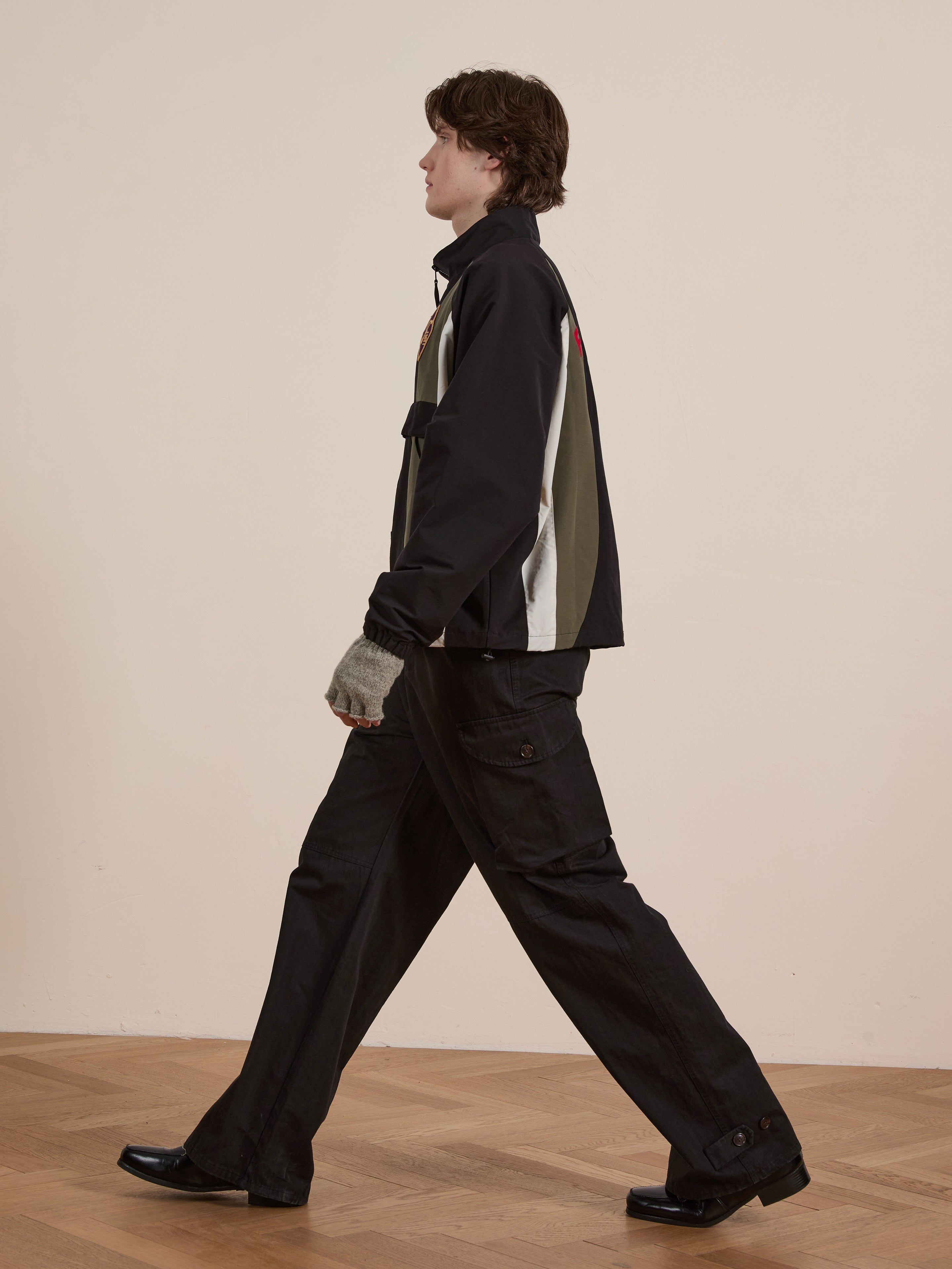 A person walking sideways in a vintage style, donning the FOUND Sporting Club Pullover Jacket in black and gray, paired with dark pants and black shoes, against a beige backdrop.