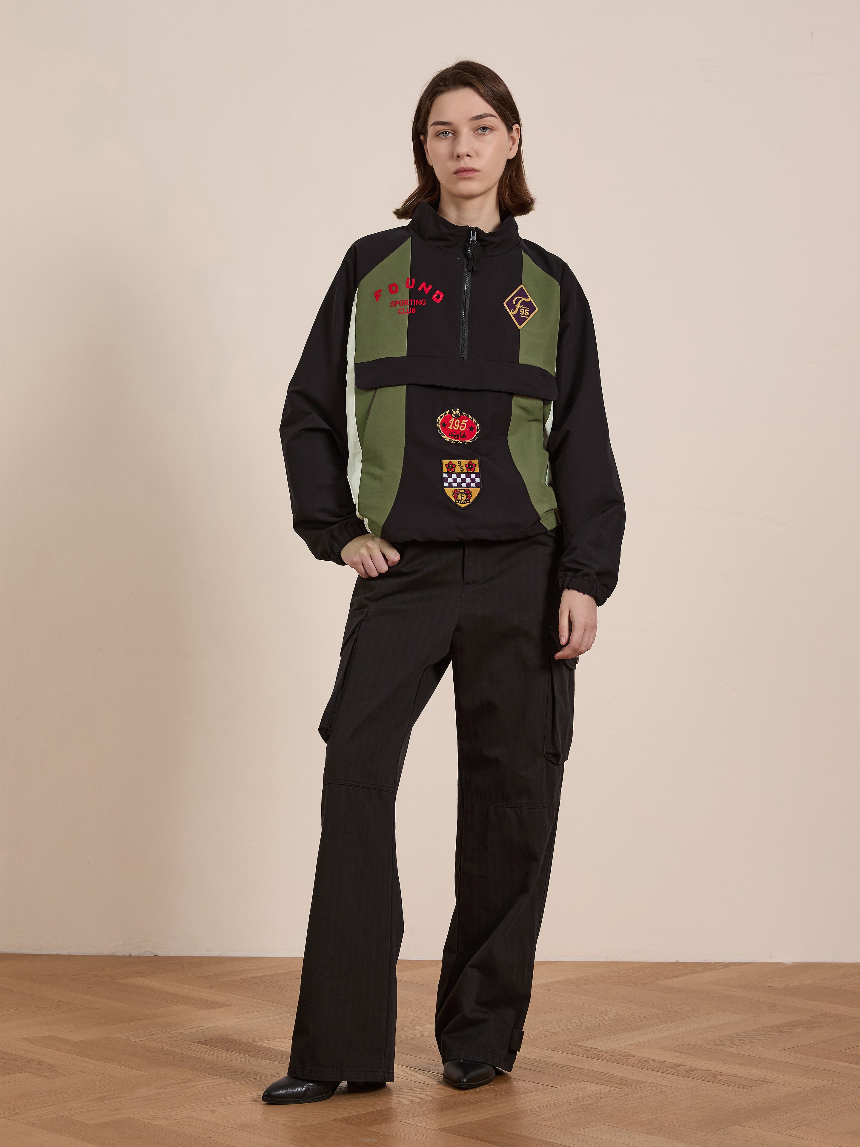 A person wearing FOUND's black and green Sporting Club Pullover Jacket with embroidered patches, black cargo pants, and black shoes stands on a wooden floor against a plain backdrop, effortlessly embodying a vintage aesthetic.