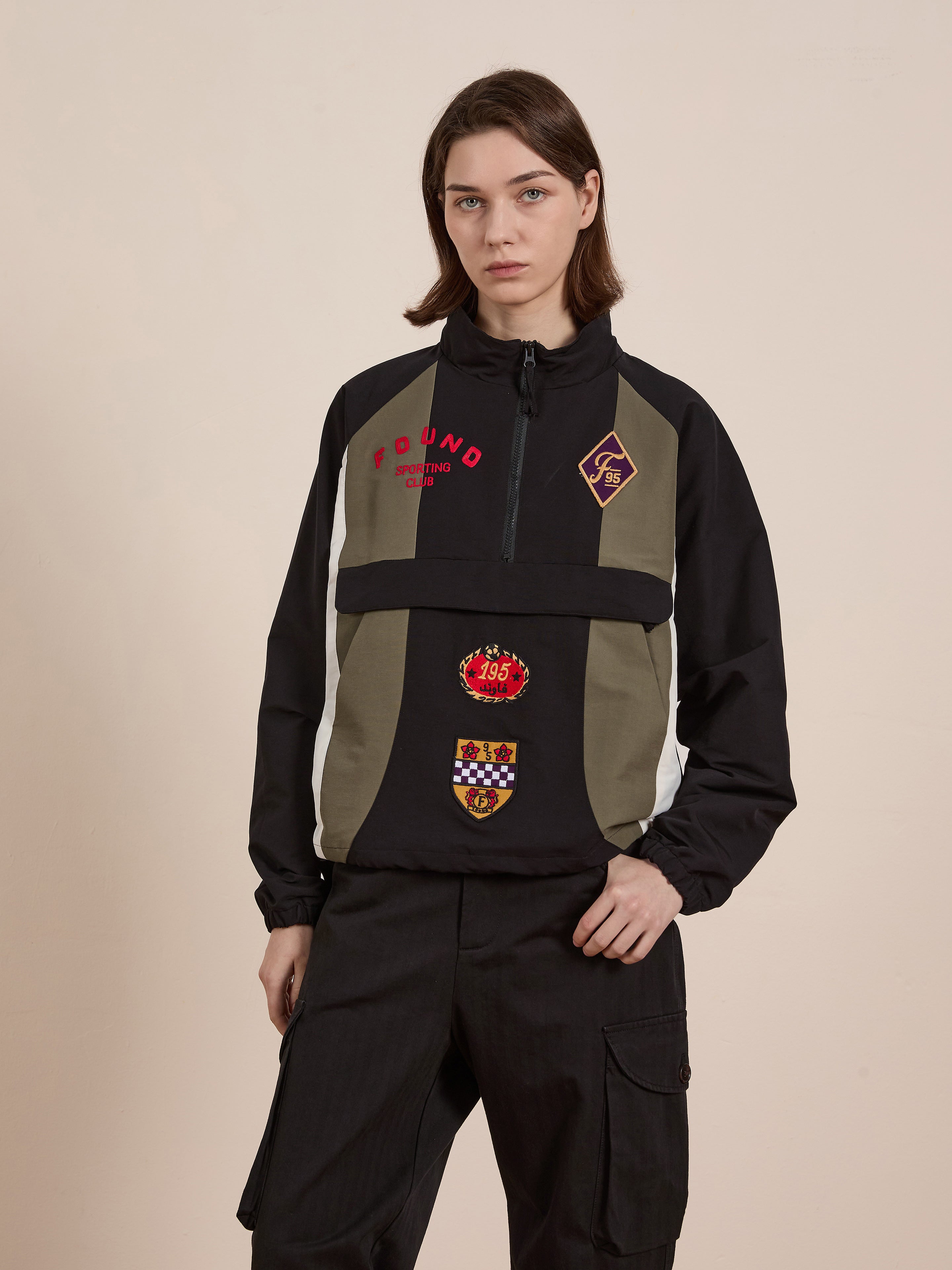 A person with shoulder-length hair stands against a plain background, wearing the FOUND Sporting Club Pullover Jacket in black and olive green, featuring a quarter-zip and multiple embroidered patches for a vintage vibe, with their hands in pockets.