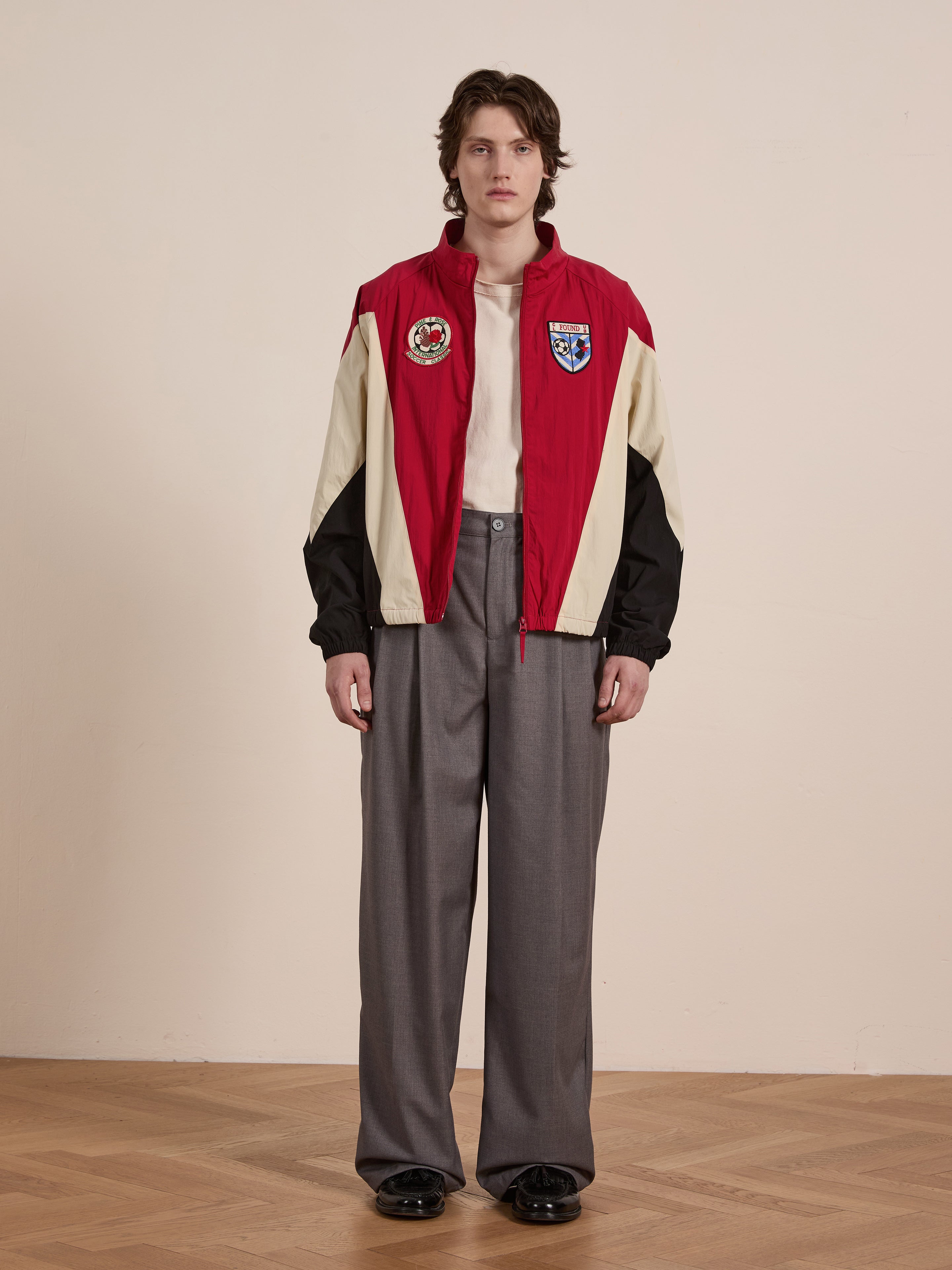 A person in FOUND's Pine and Rose Track Jacket, paired with a cream shirt and gray wide-leg trousers, stands on a wooden floor against a plain background, effortlessly combining unisex sportswear elements into their stylish look.