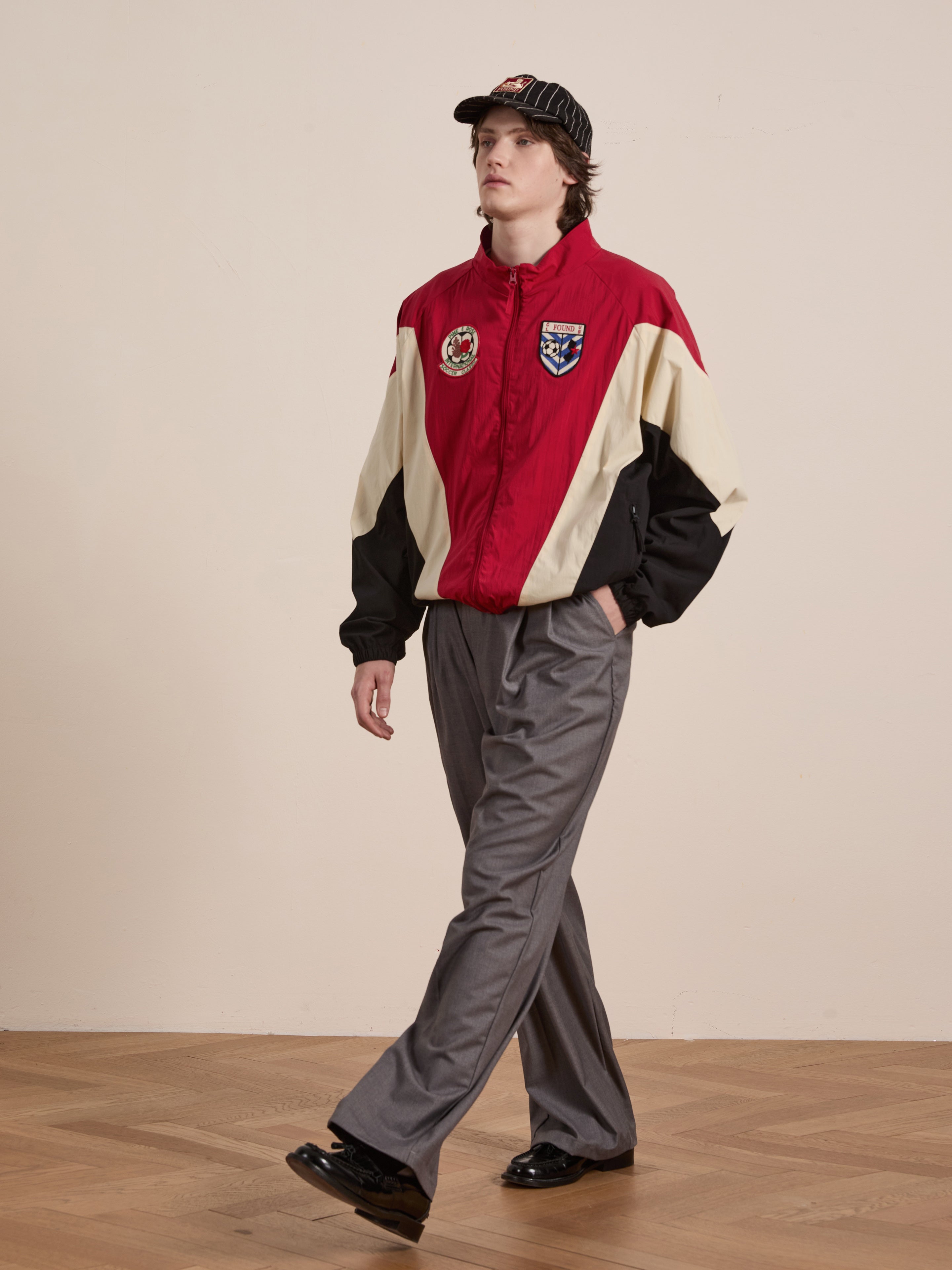 A person in a Pine and Rose Track Jacket by FOUND, gray pants, black shoes, and a black cap walks on a wooden floor against a plain background. This unisex outfit perfectly blends style and comfort for fans of versatile sportswear.
