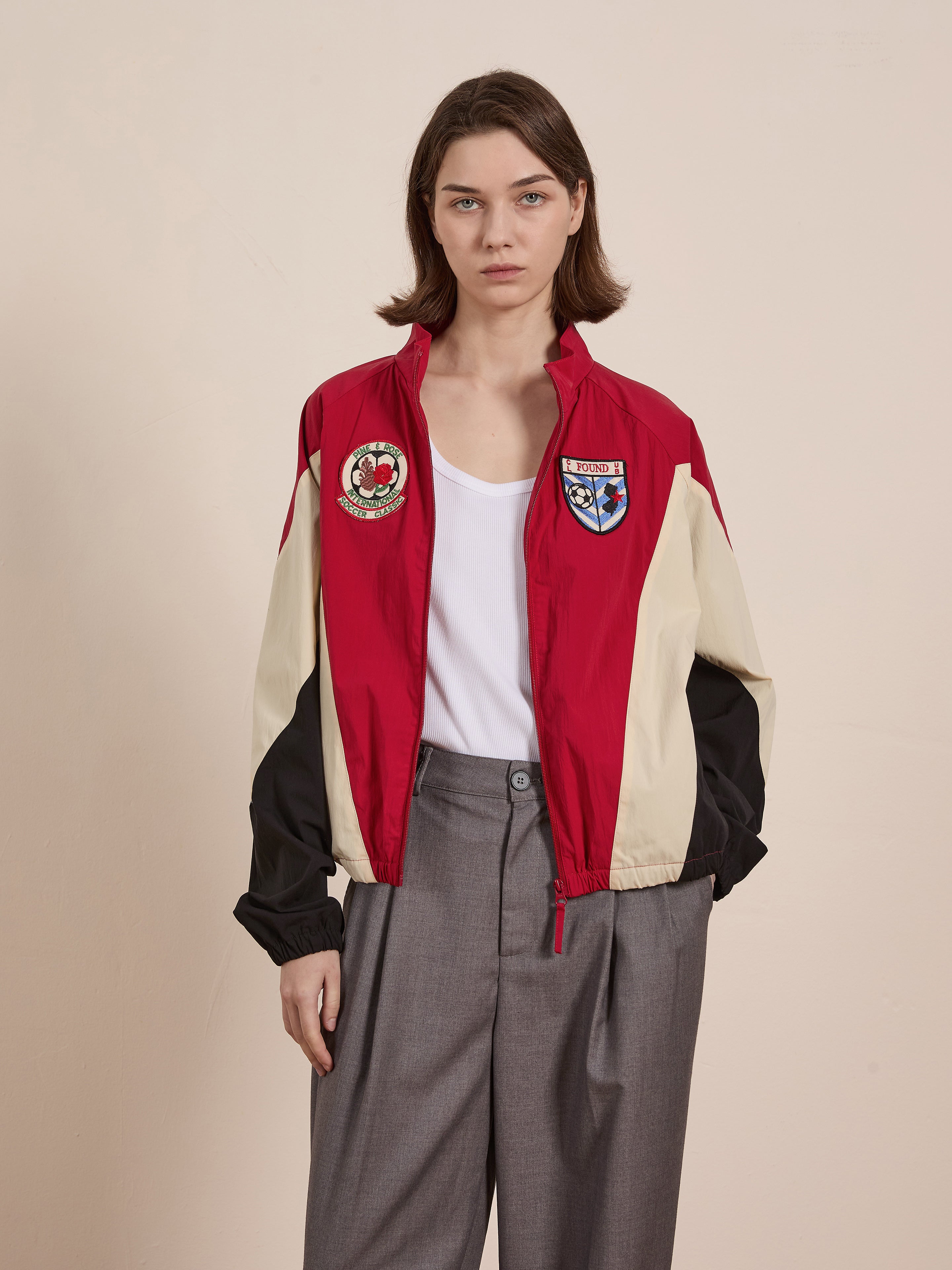 In a plain setting, an individual models the FOUND Pine and Rose Track Jacket—a unisex design in red, beige, and black with patches—accompanied by a white shirt and gray trousers.