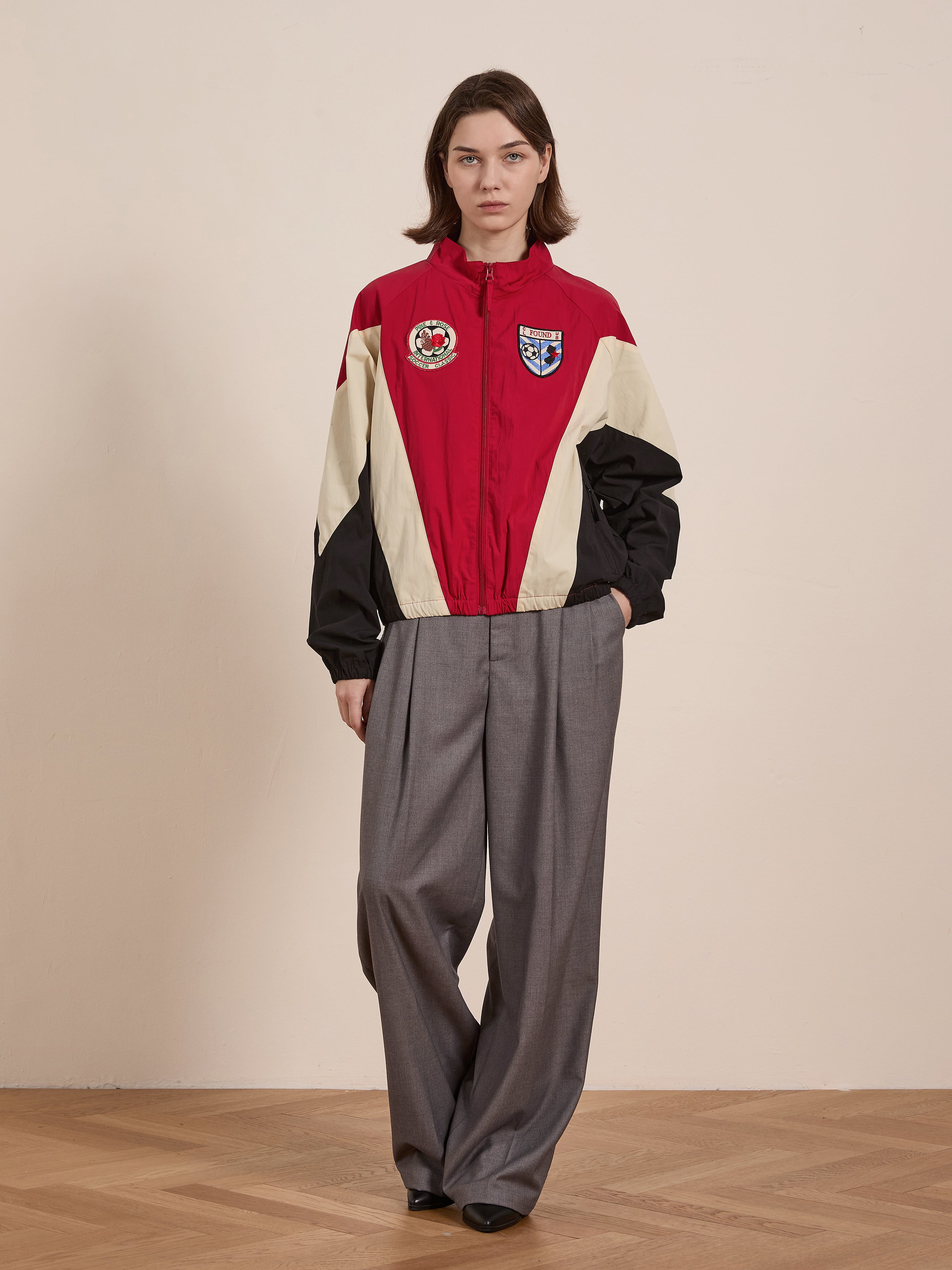 A person wearing the FOUND Pine and Rose Track Jacket with badges, paired with gray wide-leg trousers, stands on a wooden floor against a plain wall.