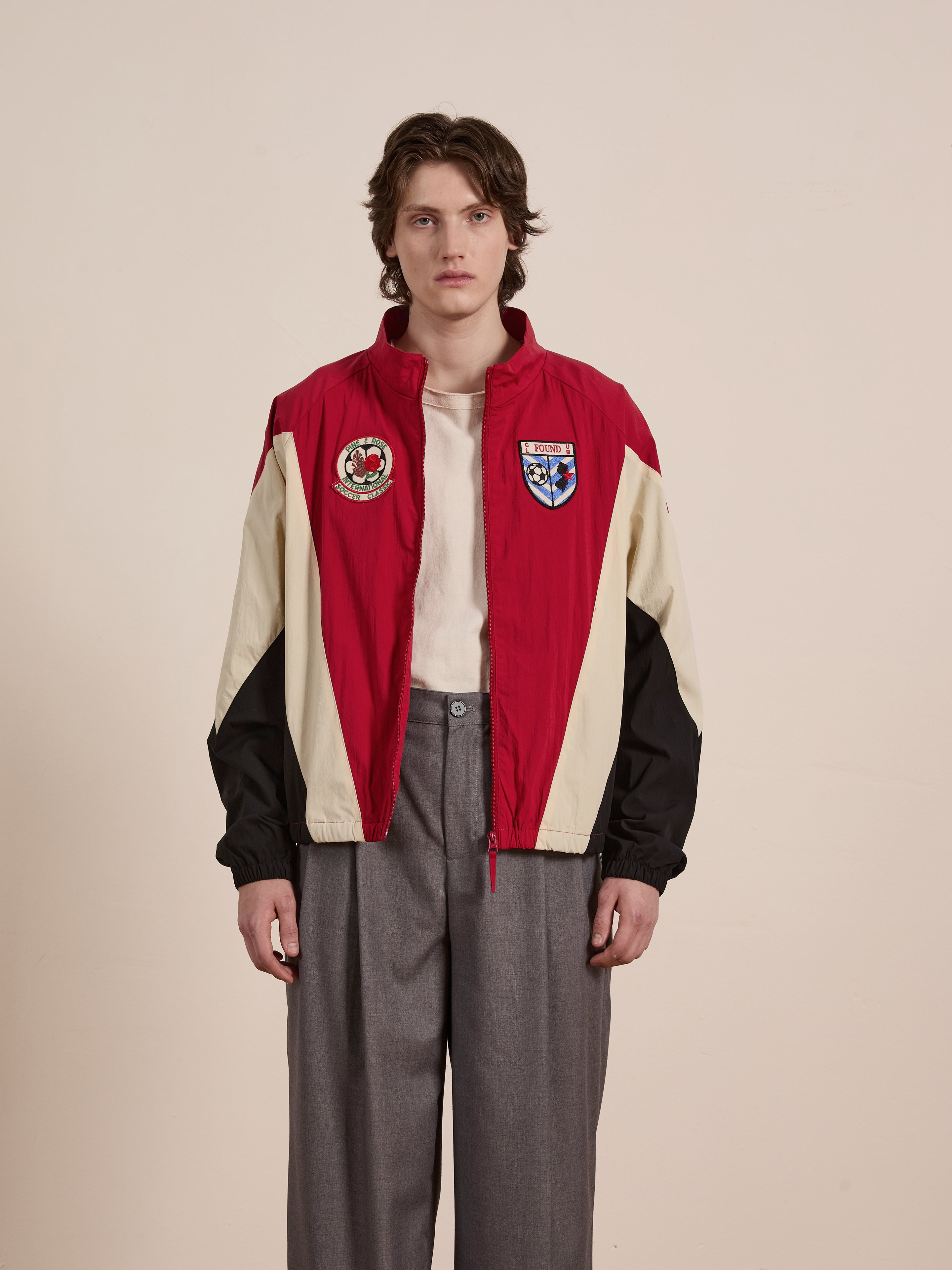 Standing against a plain background, a person wears the unisex Pine and Rose Track Jacket from FOUND in stylish red, beige, and black with patches, paired with a white shirt and gray pants. This outfit exemplifies modern sportswear fashion.