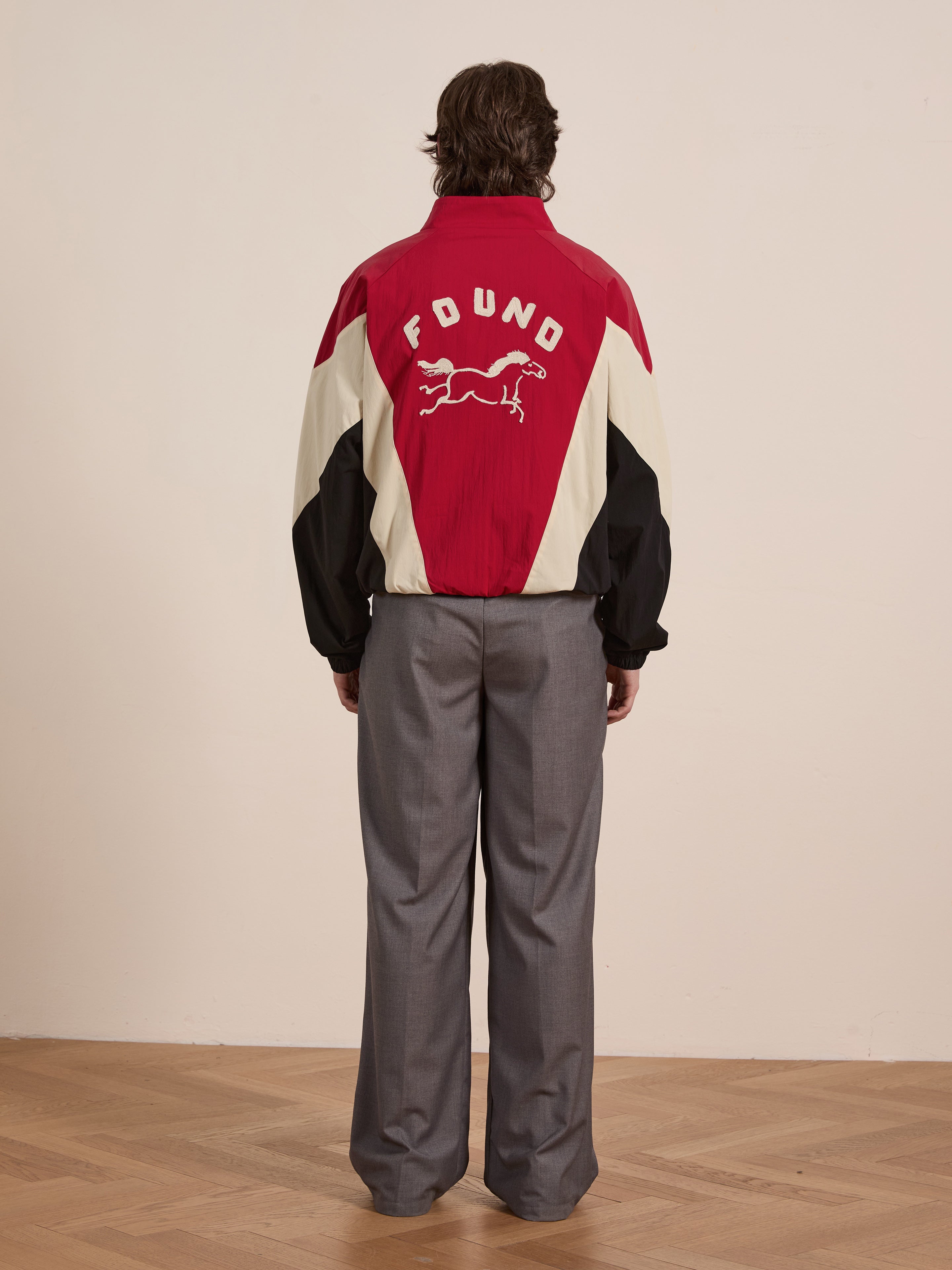 A person stands facing a wall, donning the Pine and Rose Track Jacket from FOUND, featuring red and white colors with a running animal logo on the back. It is elegantly paired with gray trousers in this unisex sportswear ensemble that blends style and comfort seamlessly.
