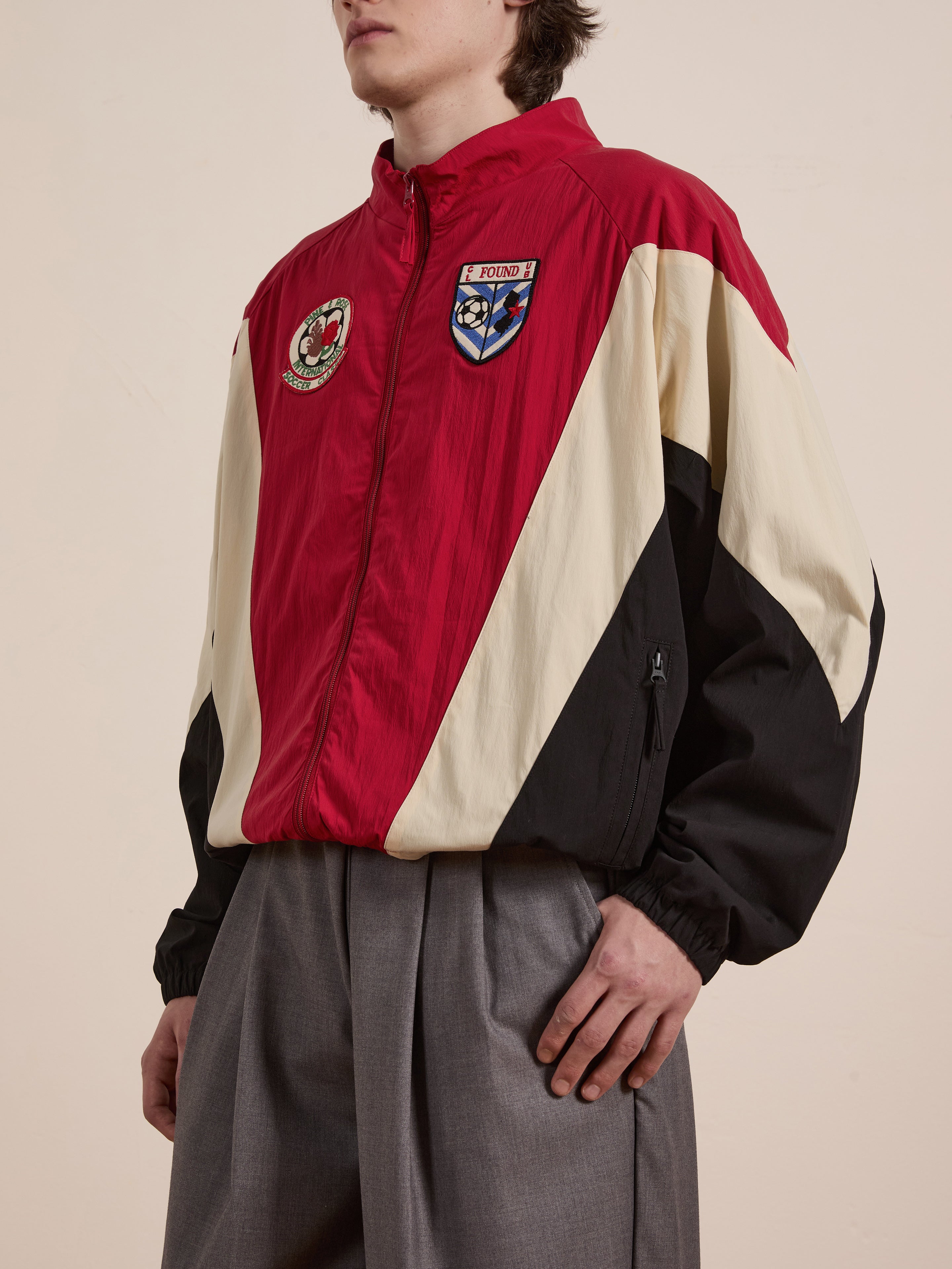 A person wears the Pine and Rose Track Jacket from FOUND, featuring stylish red, black, and cream colors with embroidered patches. Paired with gray pants, it offers a sporty yet unisex look perfect for any occasion.