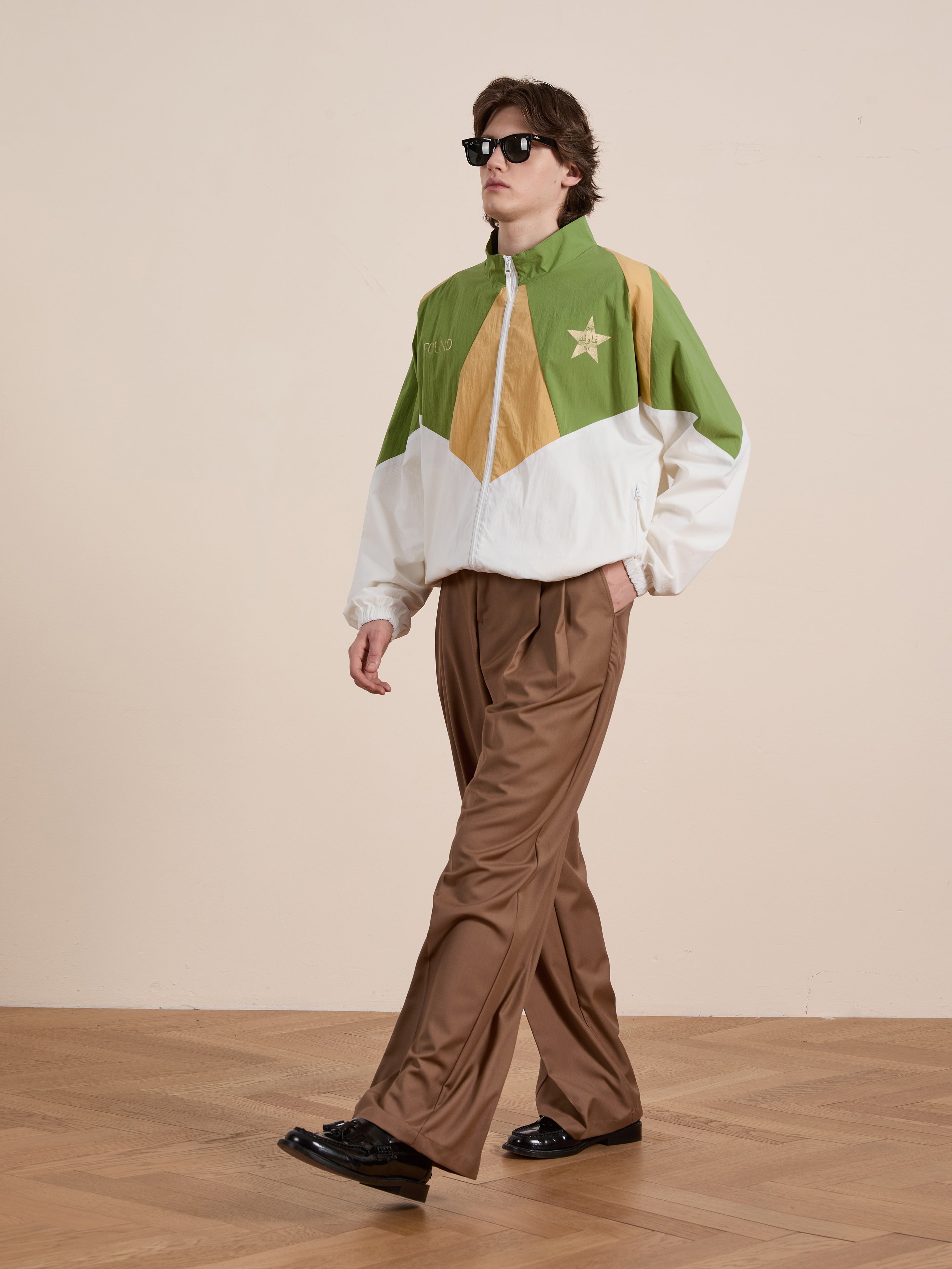 A person walking indoors is wearing sunglasses, the FOUND Legacy Cricket Track Jacket with a star, brown pants, and black shoes.
