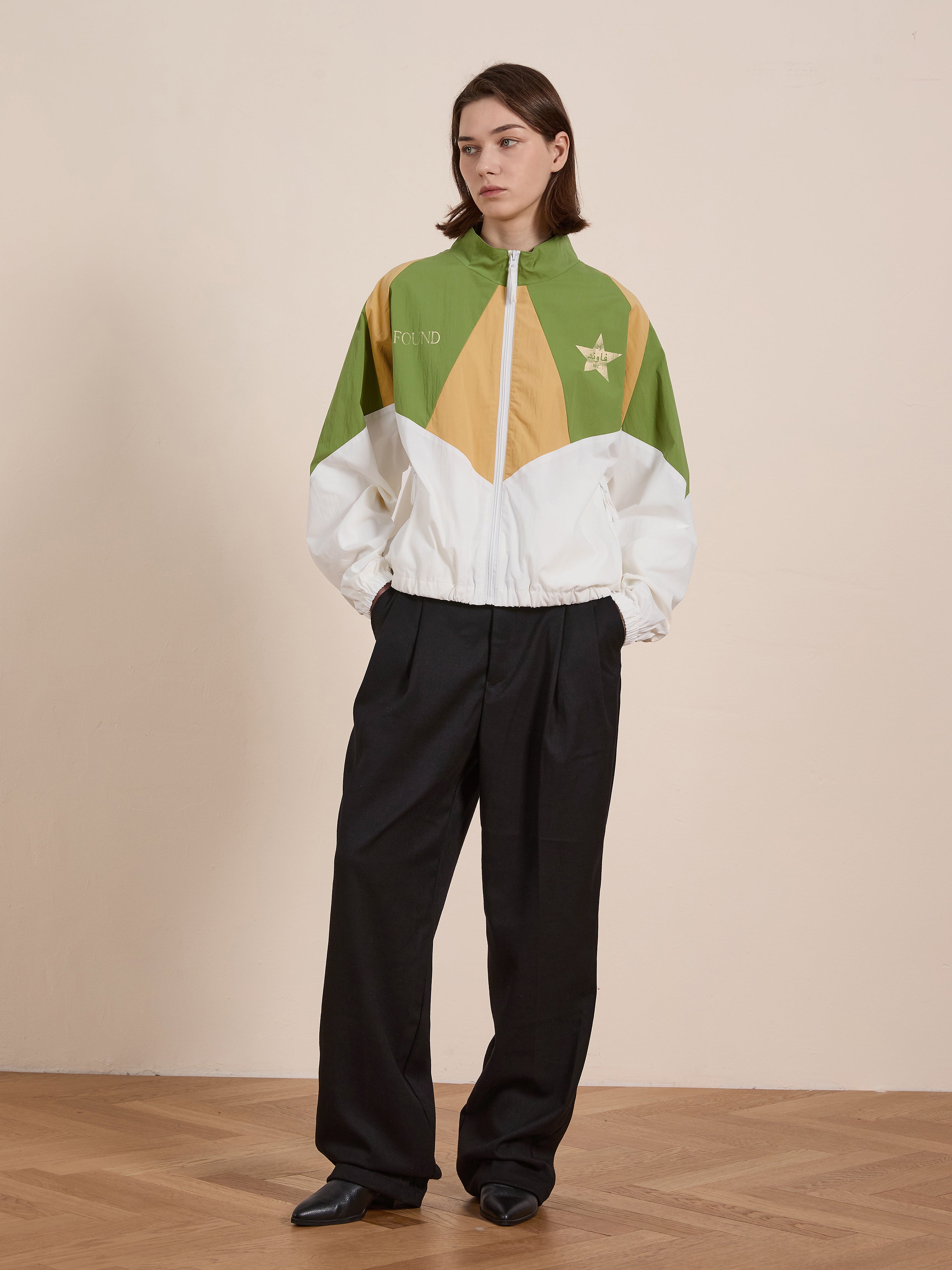 A person models FOUND's Legacy Cricket Track Jacket, showcasing green, yellow, and white vintage athletic patterns against a plain backdrop on a wooden floor. Paired with black pants, this ensemble adds a retro flair to the minimalist setting.