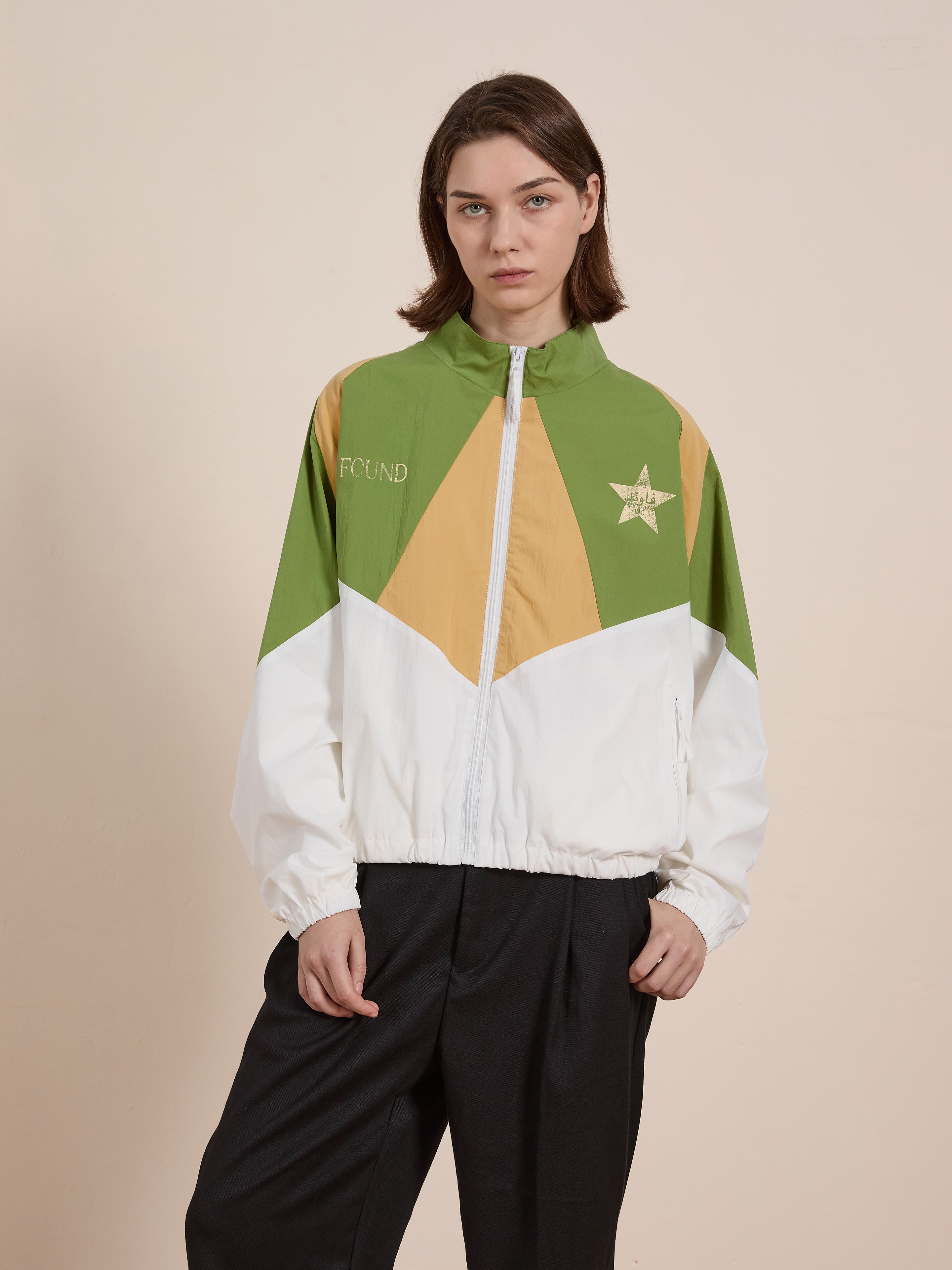 A person stands against a plain background wearing the Legacy Cricket Track Jacket by FOUND, featuring a green, white, and orange design with a star motif.
