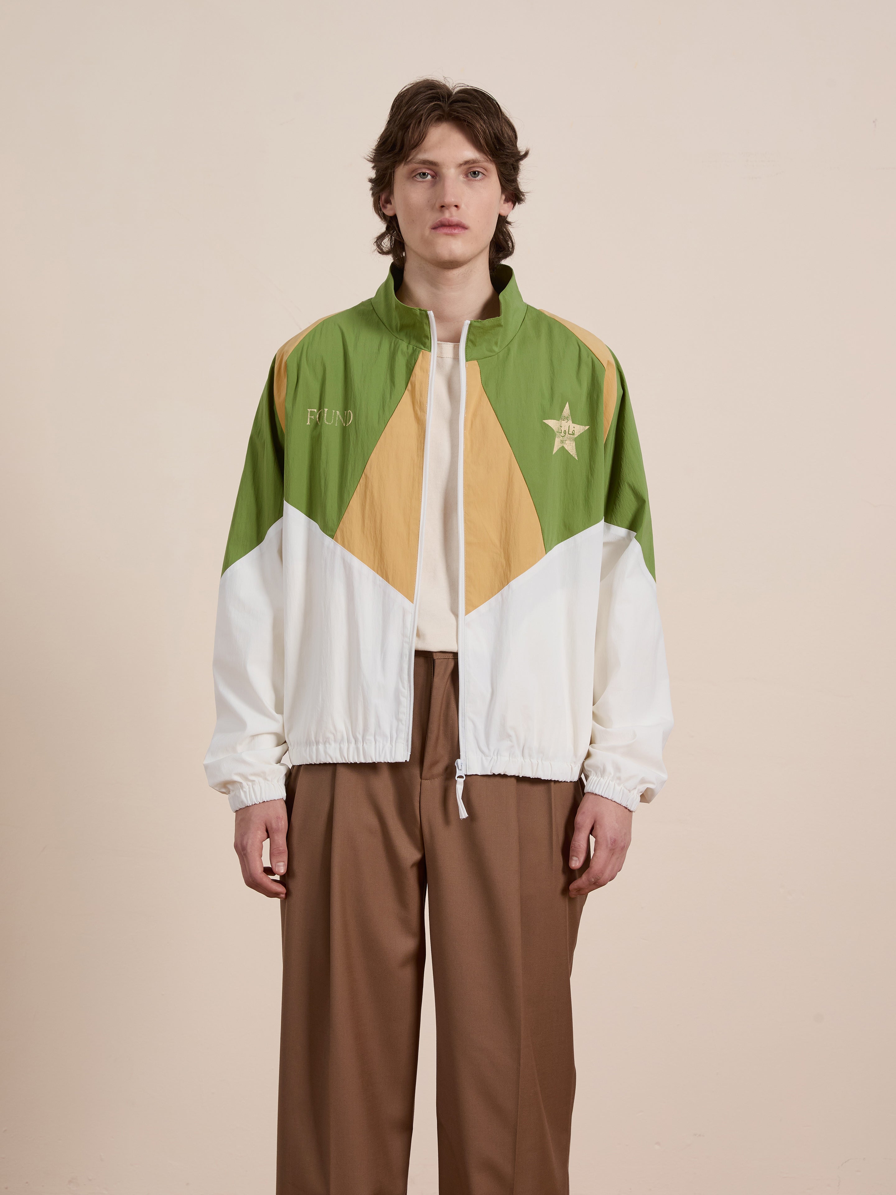 A person in the Legacy Cricket Track Jacket by FOUND, featuring green, white, and tan geometric patterns, stands against a neutral background.