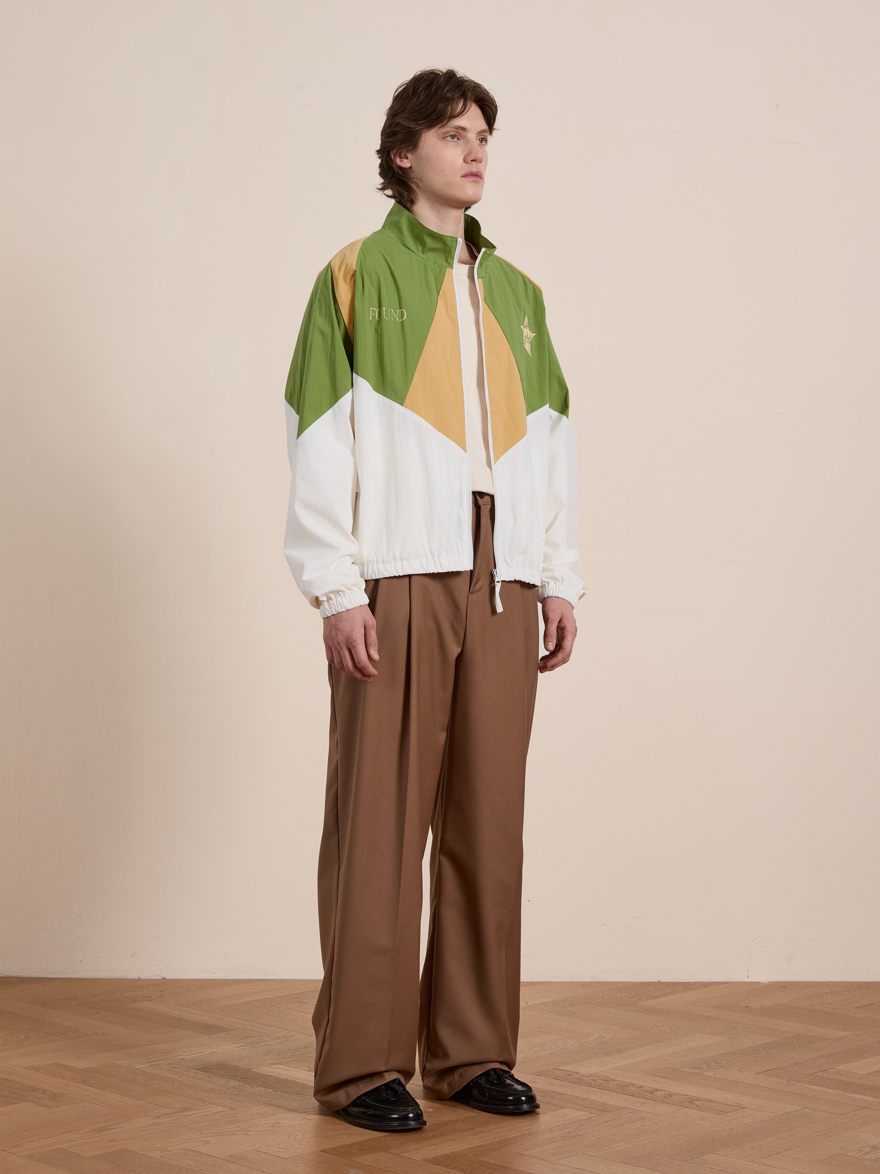 A person in a FOUND Legacy Cricket Track Jacket, featuring green, yellow, and white colors, stands on a wooden floor against a plain wall. They pair it with brown pants and black shoes.
