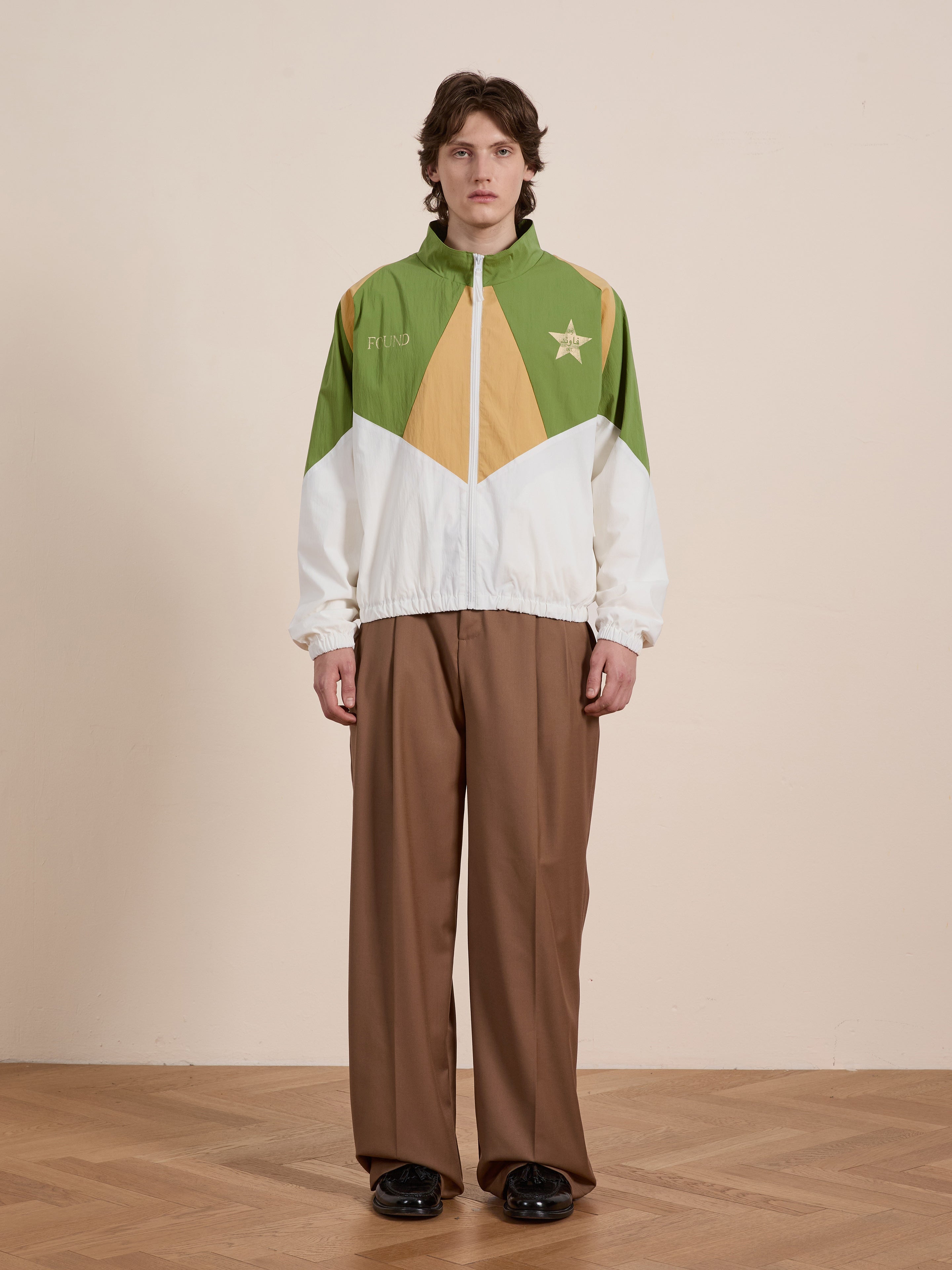 A person is wearing a Legacy Cricket Track Jacket by FOUND, featuring green, white, and yellow hues. They have paired it with wide brown pants and black shoes, standing on a wooden floor in front of a plain wall.