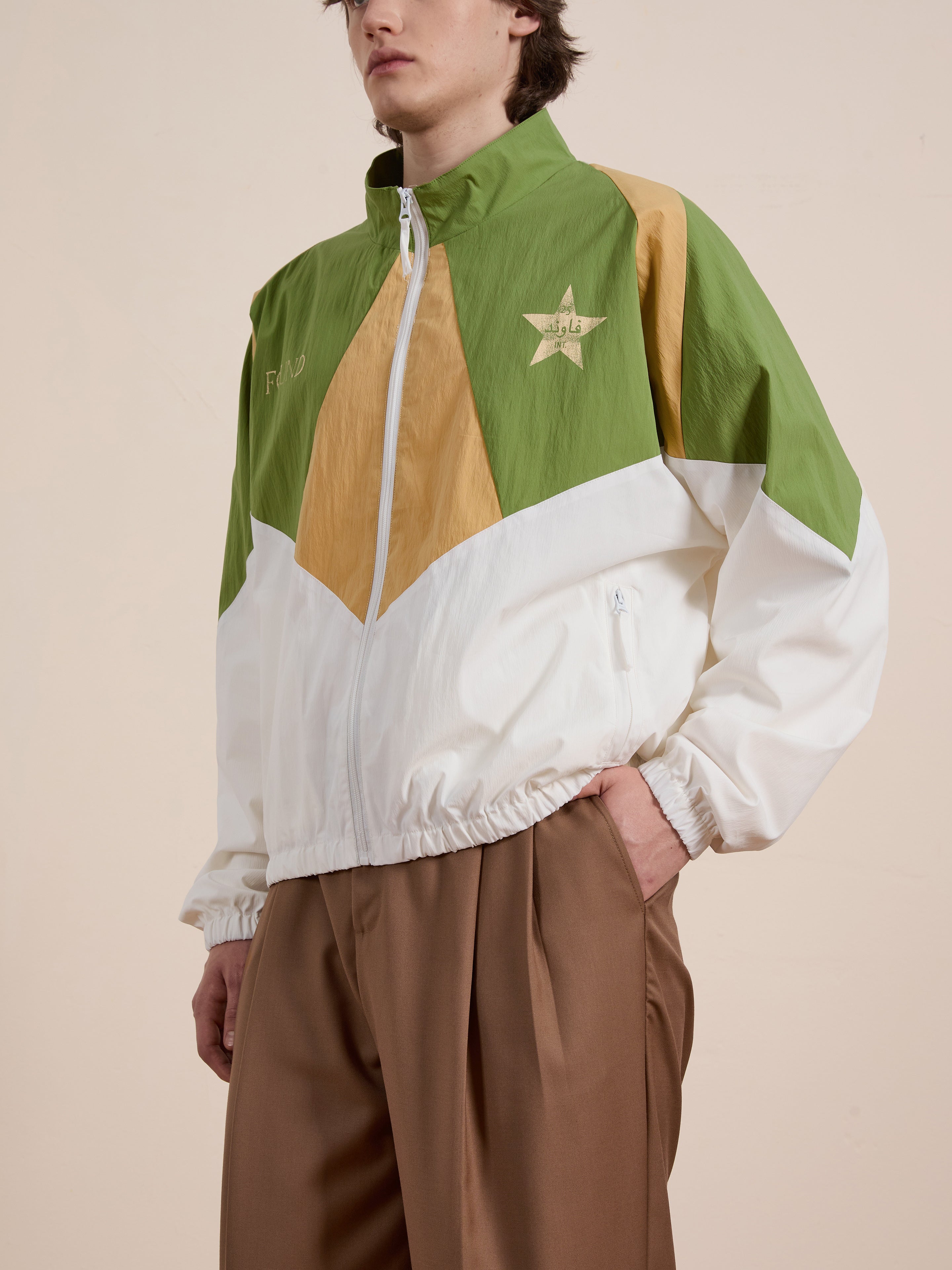 A person is wearing FOUND's Legacy Cricket Track Jacket in green, white, and beige with a star emblem, paired with brown pants. The background is plain.