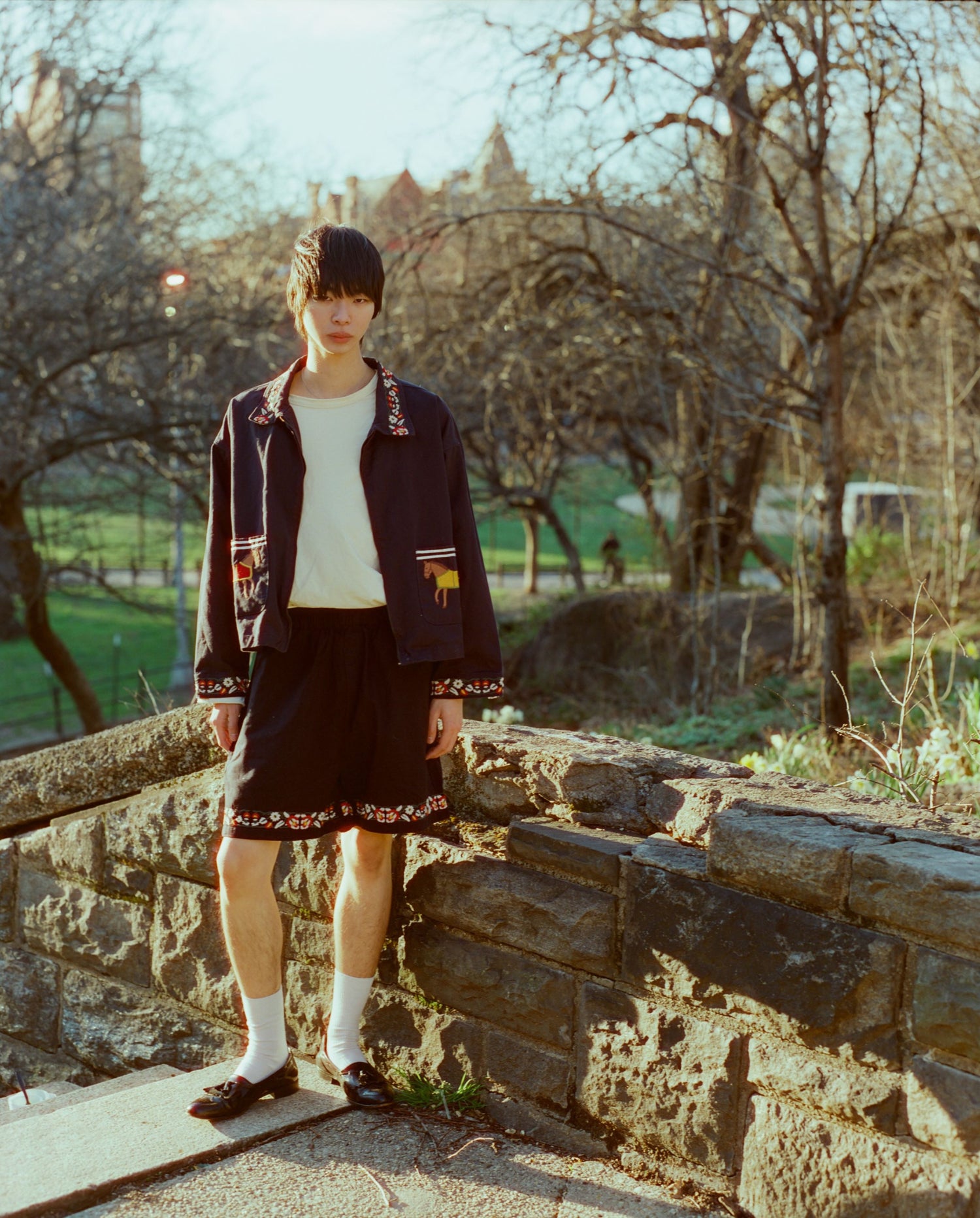 Young jockey in racing colours standing on a stone wall with trees and a building in the background, during sunset wearing Found's Horse Equine Work Jacket.
