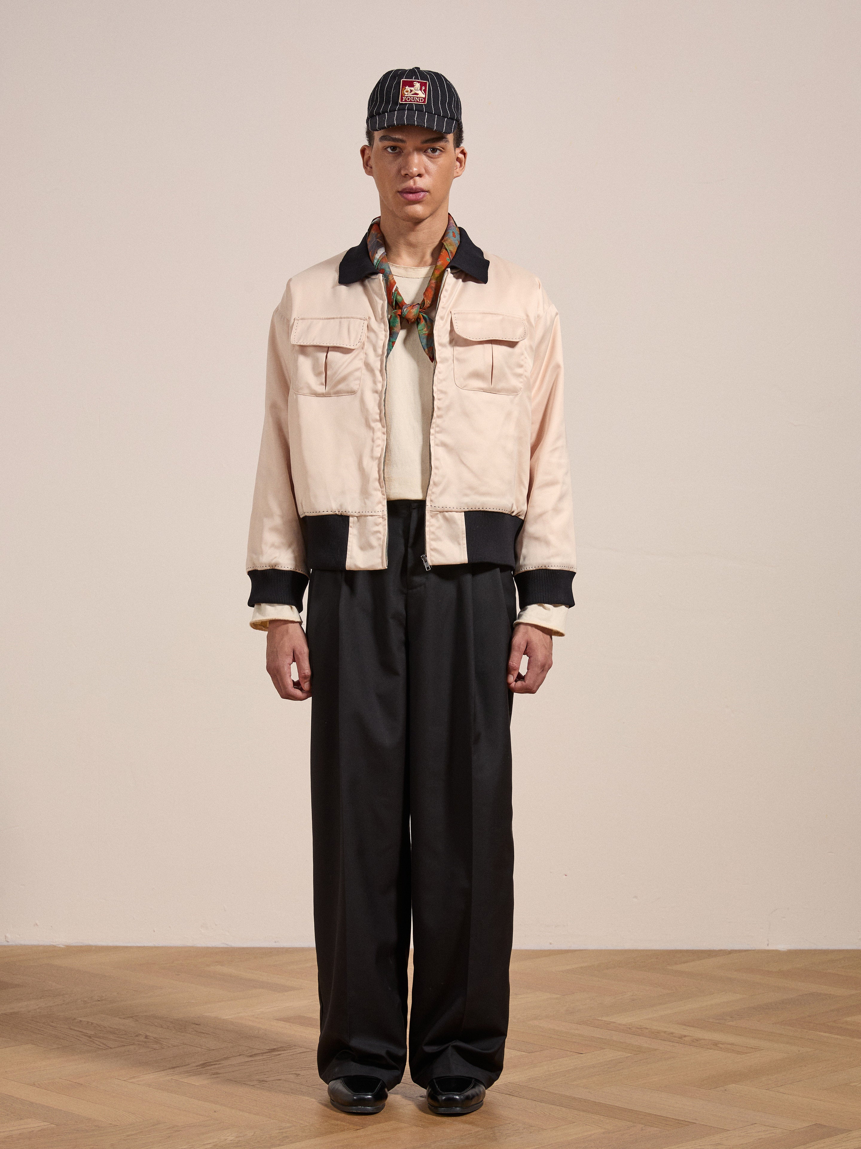 A person stands on a wooden floor, wearing the Daman Satin Jacket from FOUND, dark pants, a cap, and a scarf around their neck. They have a neutral expression with their hands at their sides.