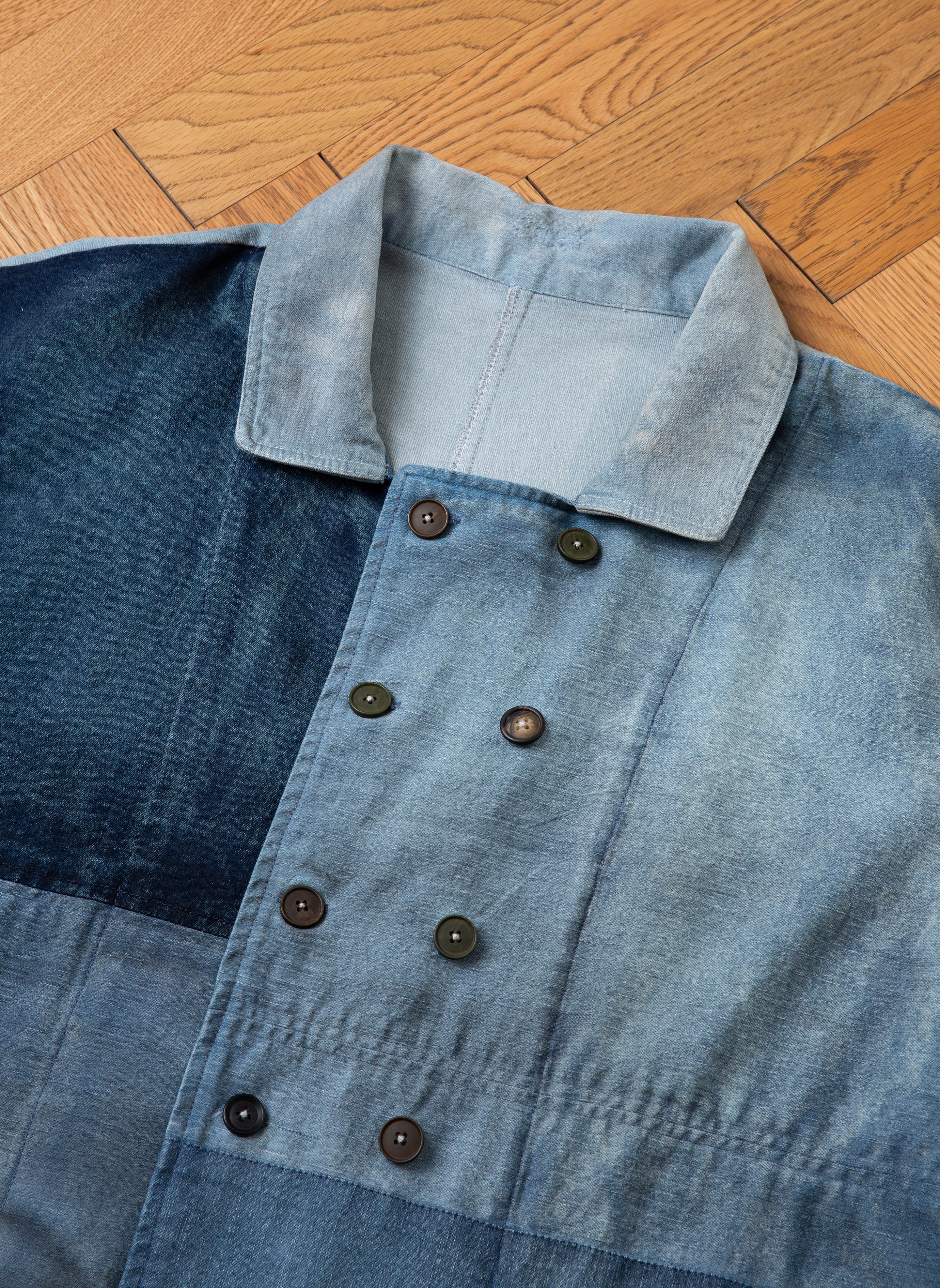 A close-up of the Multi Patch Painters Work Jacket by FOUND, showcasing its blue denim fabric with a patchwork design and hand distressing, featuring a double-breasted button arrangement, laid flat on a wooden floor.