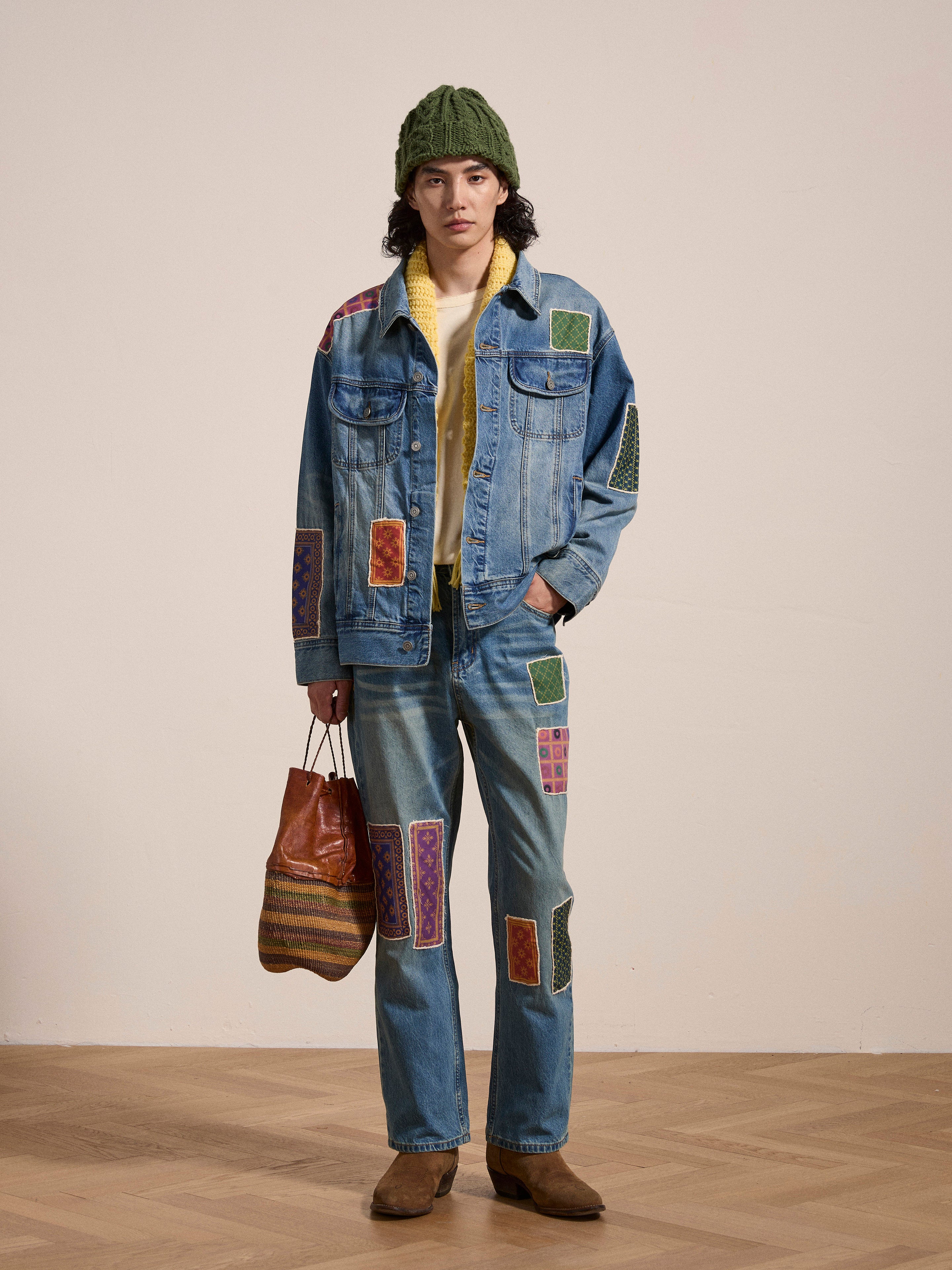 A person wearing a denim jacket and the Kantha Patchwork Jeans by Found, featuring South Asian inspired patches, paired with a green knit hat, brown shoes, and holding a striped tote bag. The vintage blue wash on the jeans adds an extra layer of uniqueness to the ensemble.