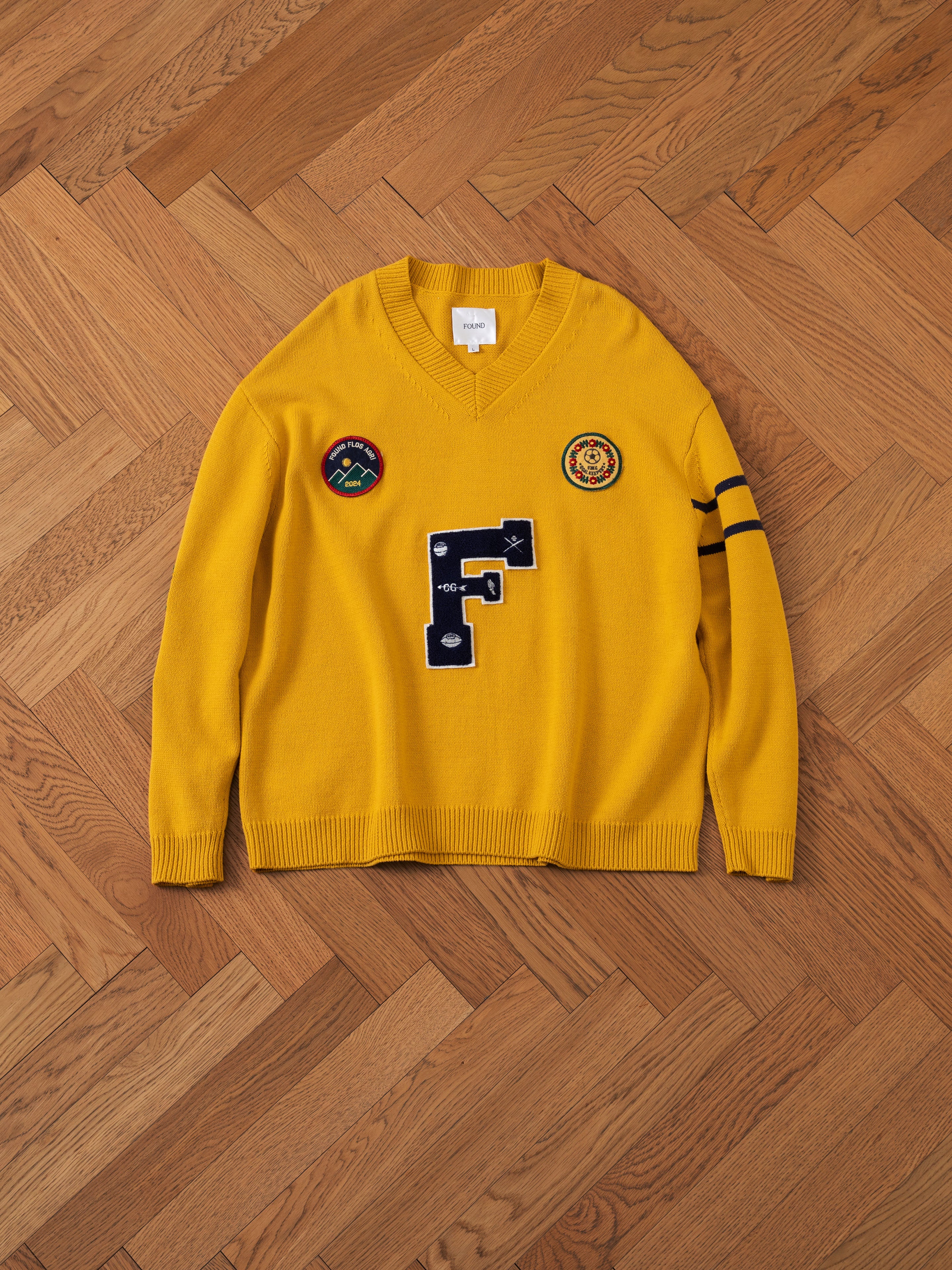 The Arrow-Wood Varsity Knit Sweater by FOUND is a yellow V-neck sweater adorned with embroidered patches and designs, including a prominent blue letter "F" on the front. Displayed on a wooden herringbone floor, it's the ideal choice for adding a vintage flair to your Ivy League wardrobe.