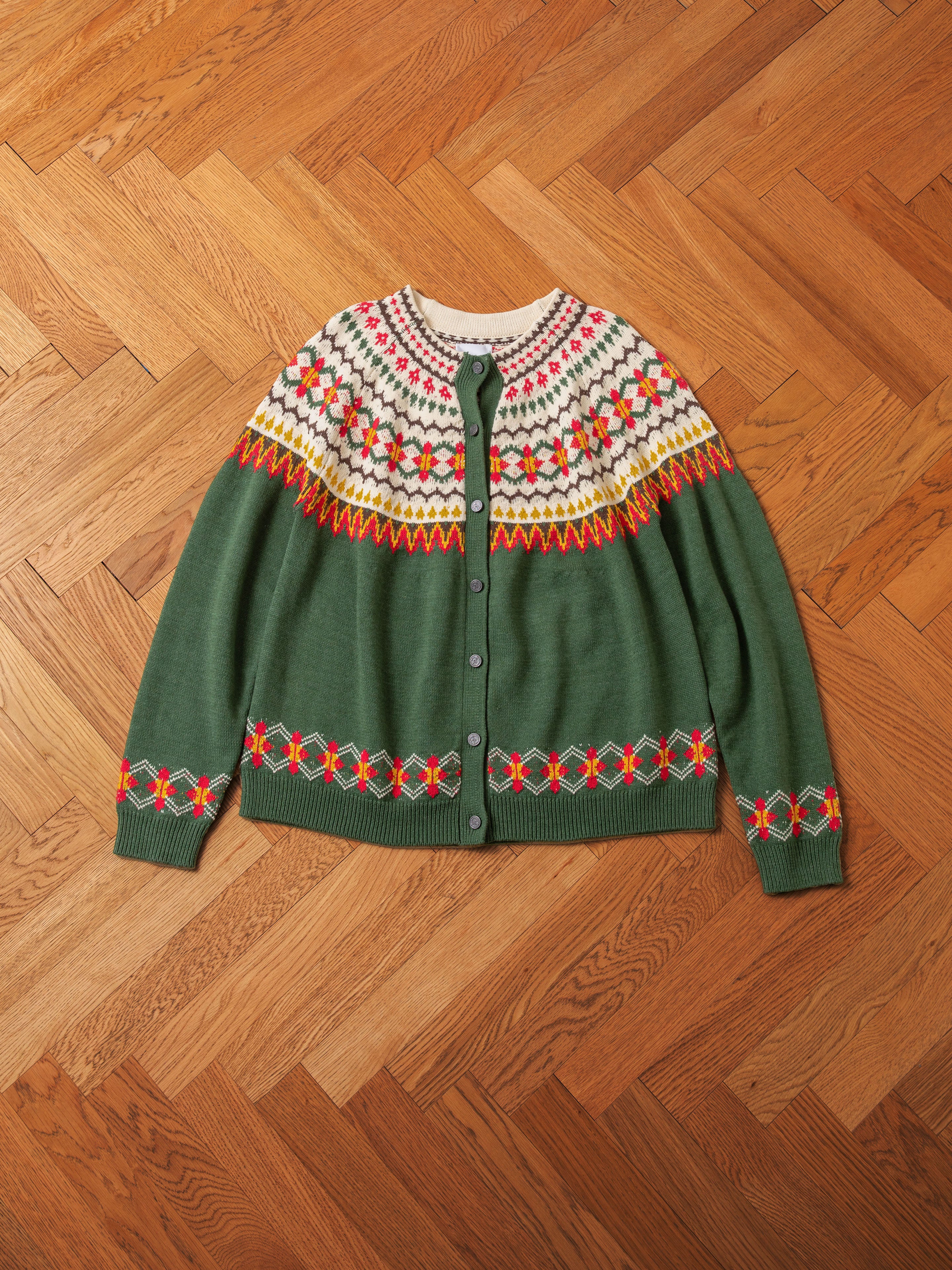 The Jahan Isles Knit Cardigan by FOUND, featuring an oversized fit and antique silver buttons, showcases a green button-up design with a multicolored, intricate knitted pattern around the collar and sleeves. It is laid flat on a wooden herringbone floor.